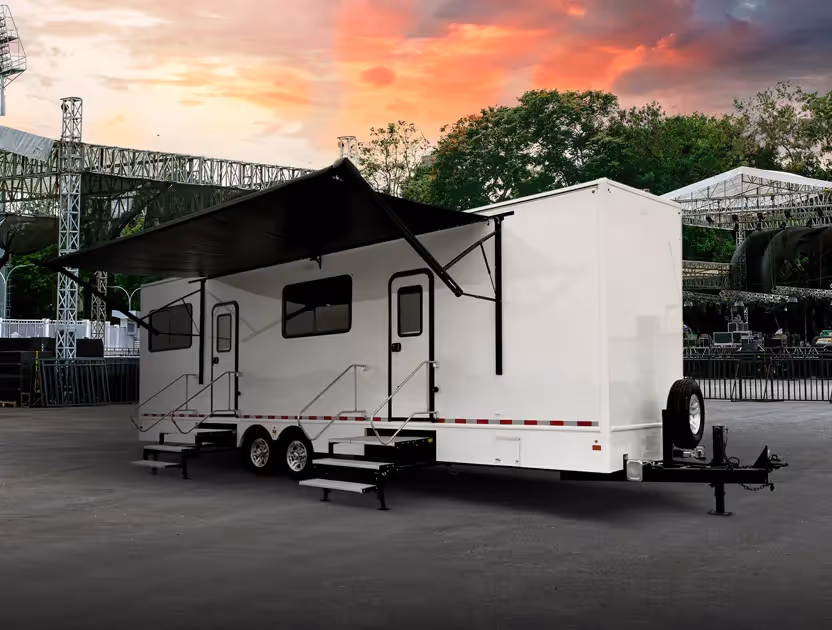 White trailer with two doors and an extended black awning parked near stage structures at sunset.