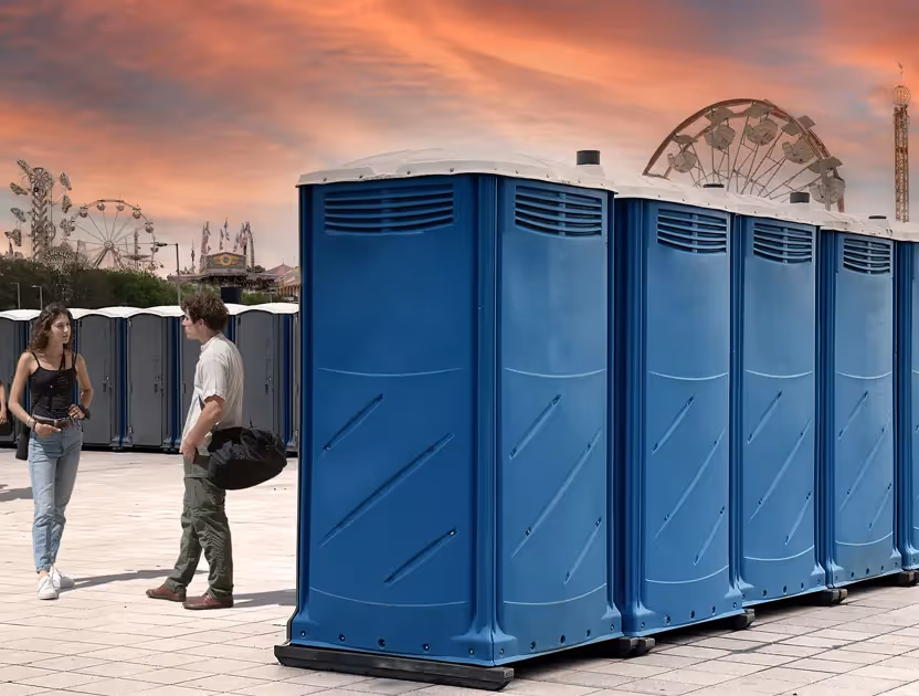 Two people standing near a row of blue portable toilets with a sunset sky and carnival rides in background.