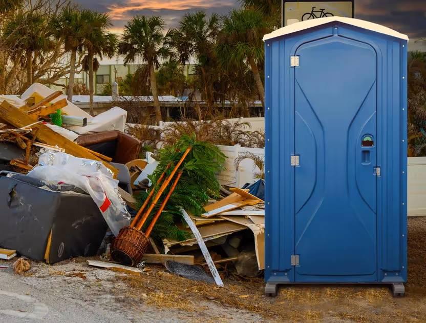 Blue portable toilet next to a pile of wood debris and broken furniture outdoors at sunset.