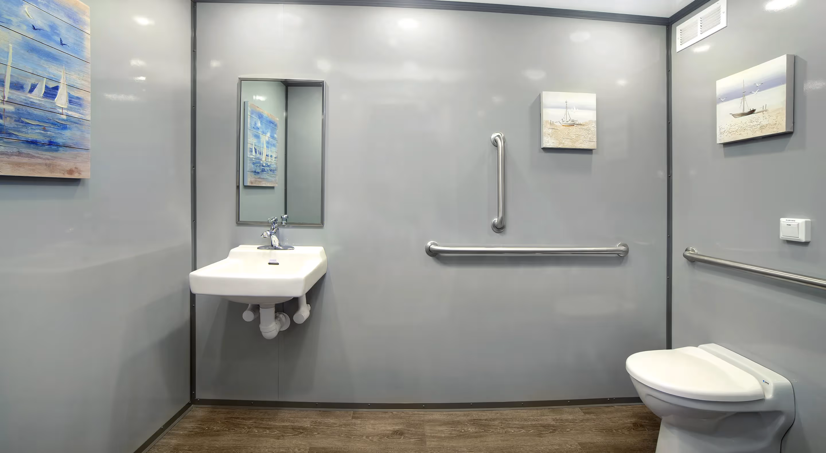Accessible bathroom with wall-mounted sink, grab bars, toilet, nautical paintings, and wood-look floor.