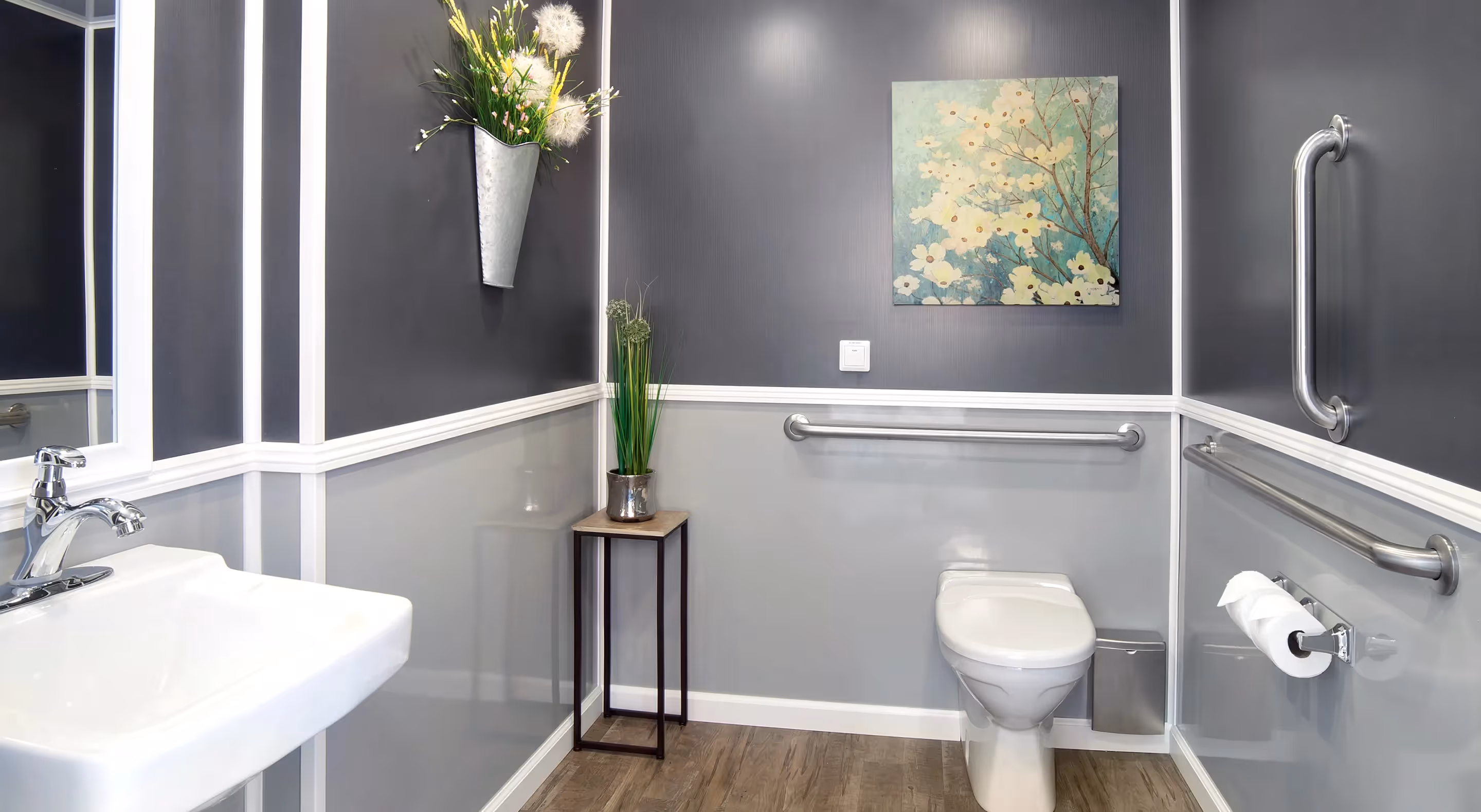 Modern accessible bathroom with sink, toilet, steel grab bars, and floral artwork on gray walls.