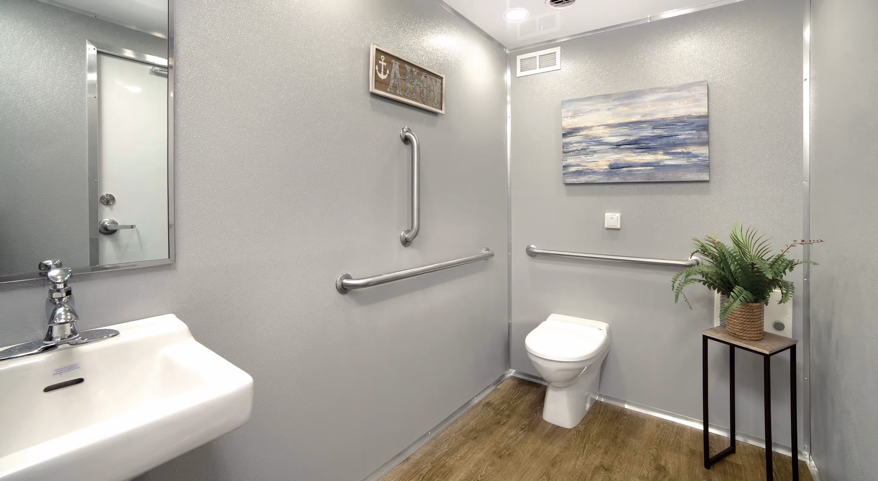 Modern accessible restroom with sink, toilet, grab bars, ocean painting, and small plant on stand.