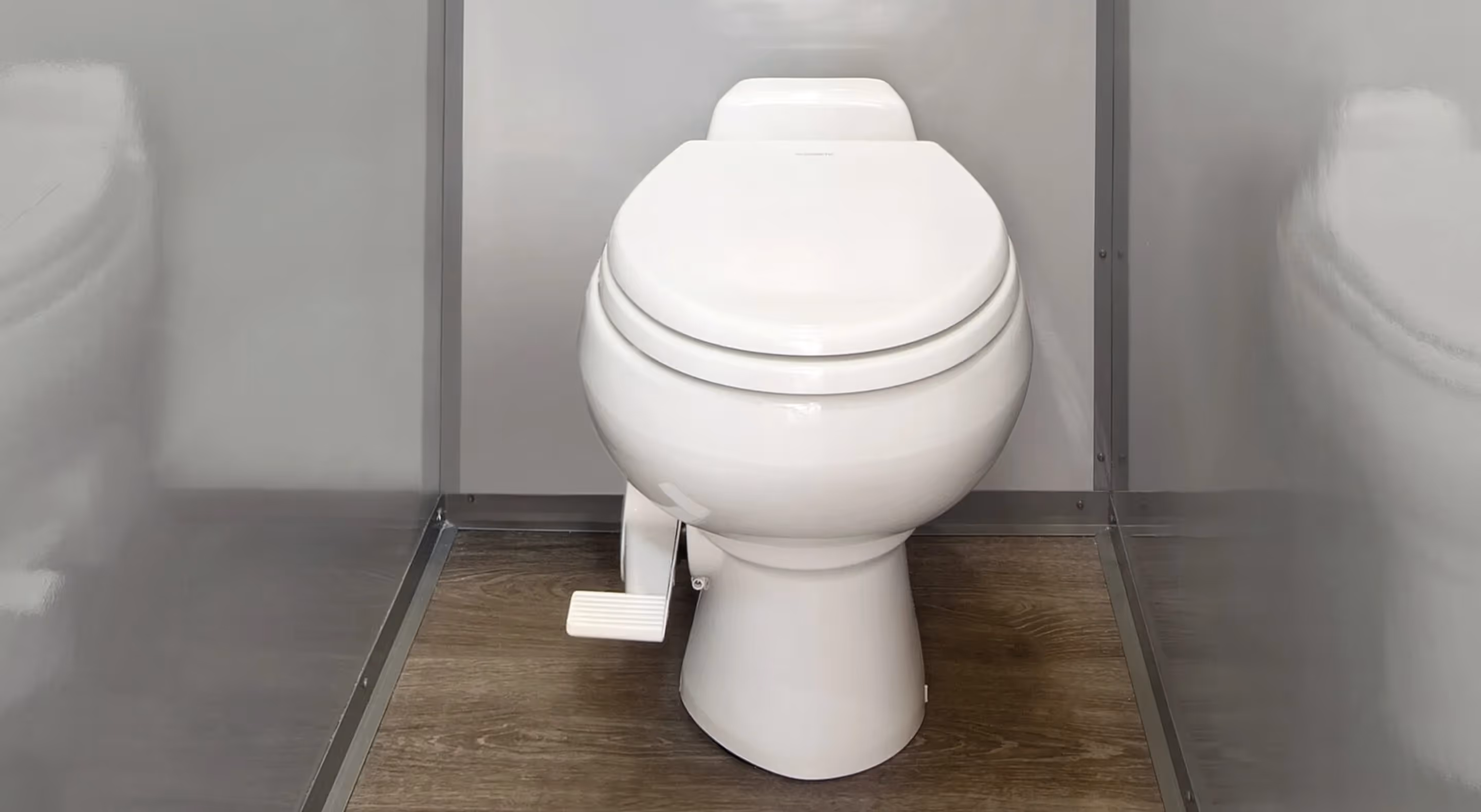 White marine-style toilet with foot pedal inside a compartment with gray walls and wood floor.