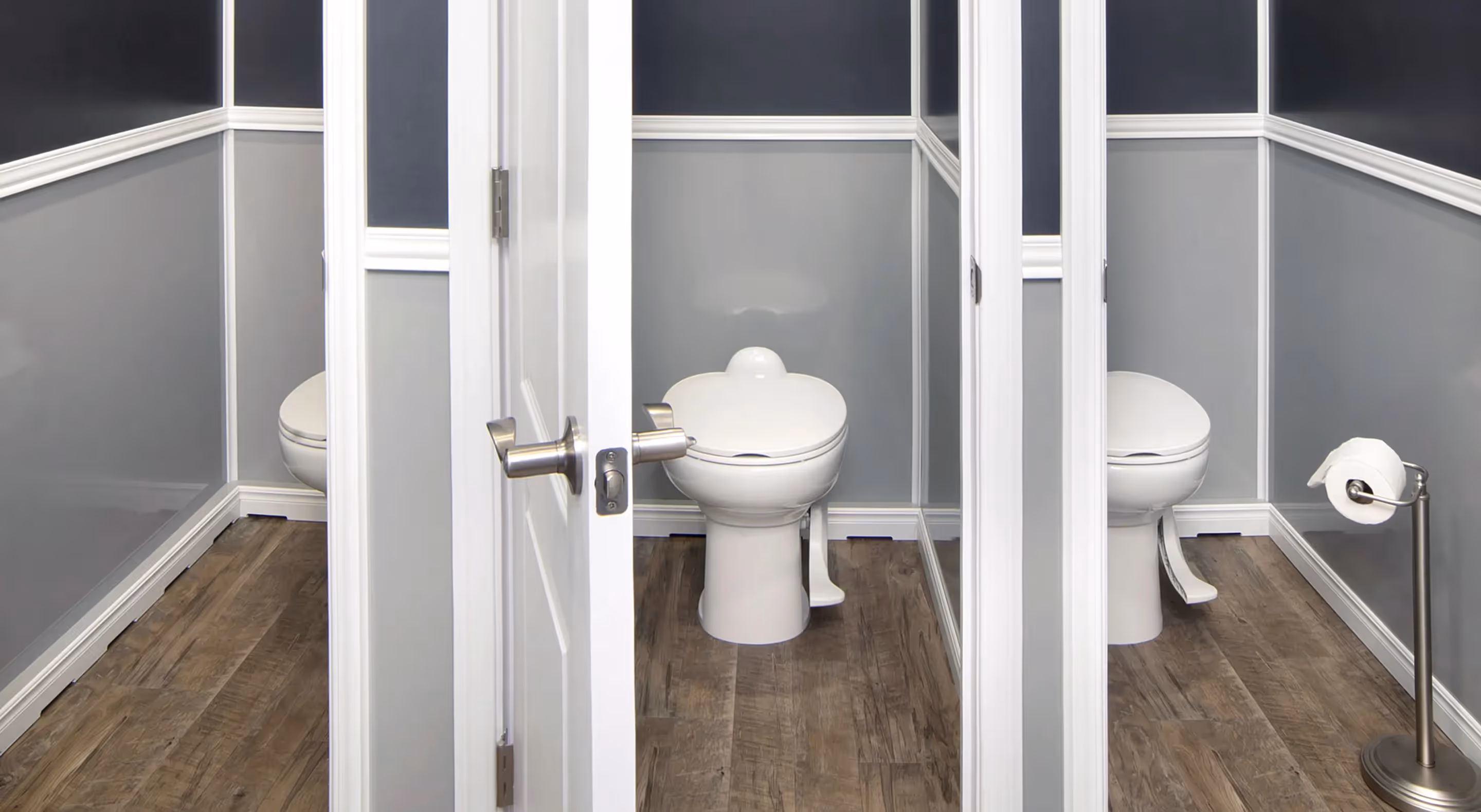 Three white toilets in separate stalls with gray walls and wood floors, one with a toilet paper stand.
