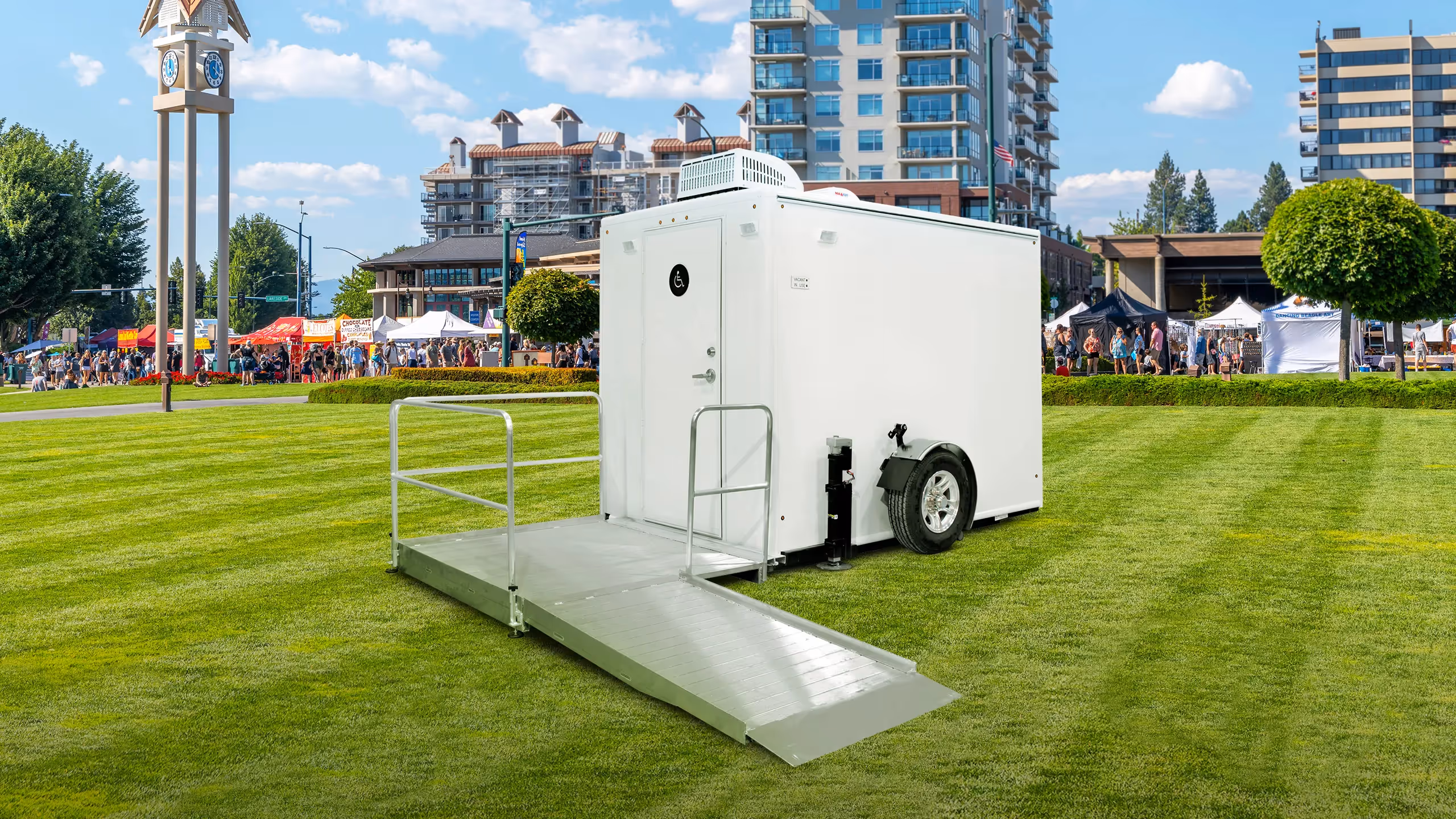 ADA-compliant 1-station restroom trailer on grass, surrounded by buildings and a festival.