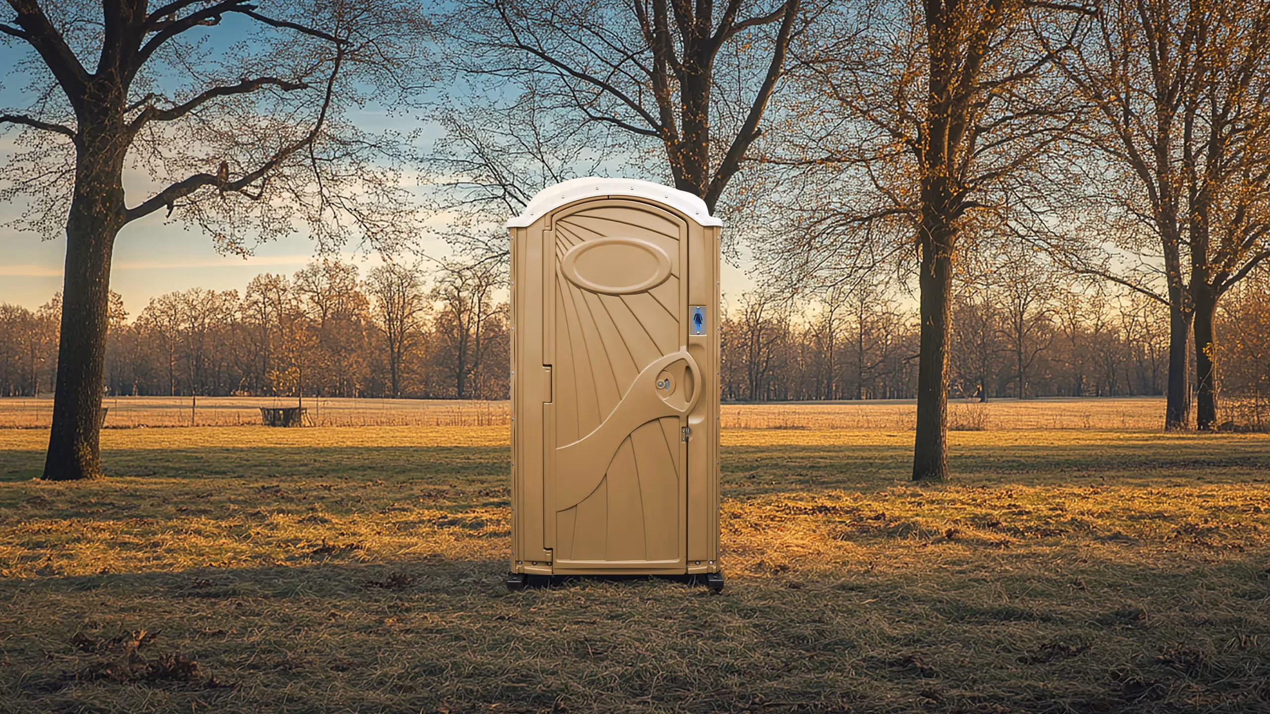 Portable beige outdoor restroom standing on grass in a field with bare trees at sunset.