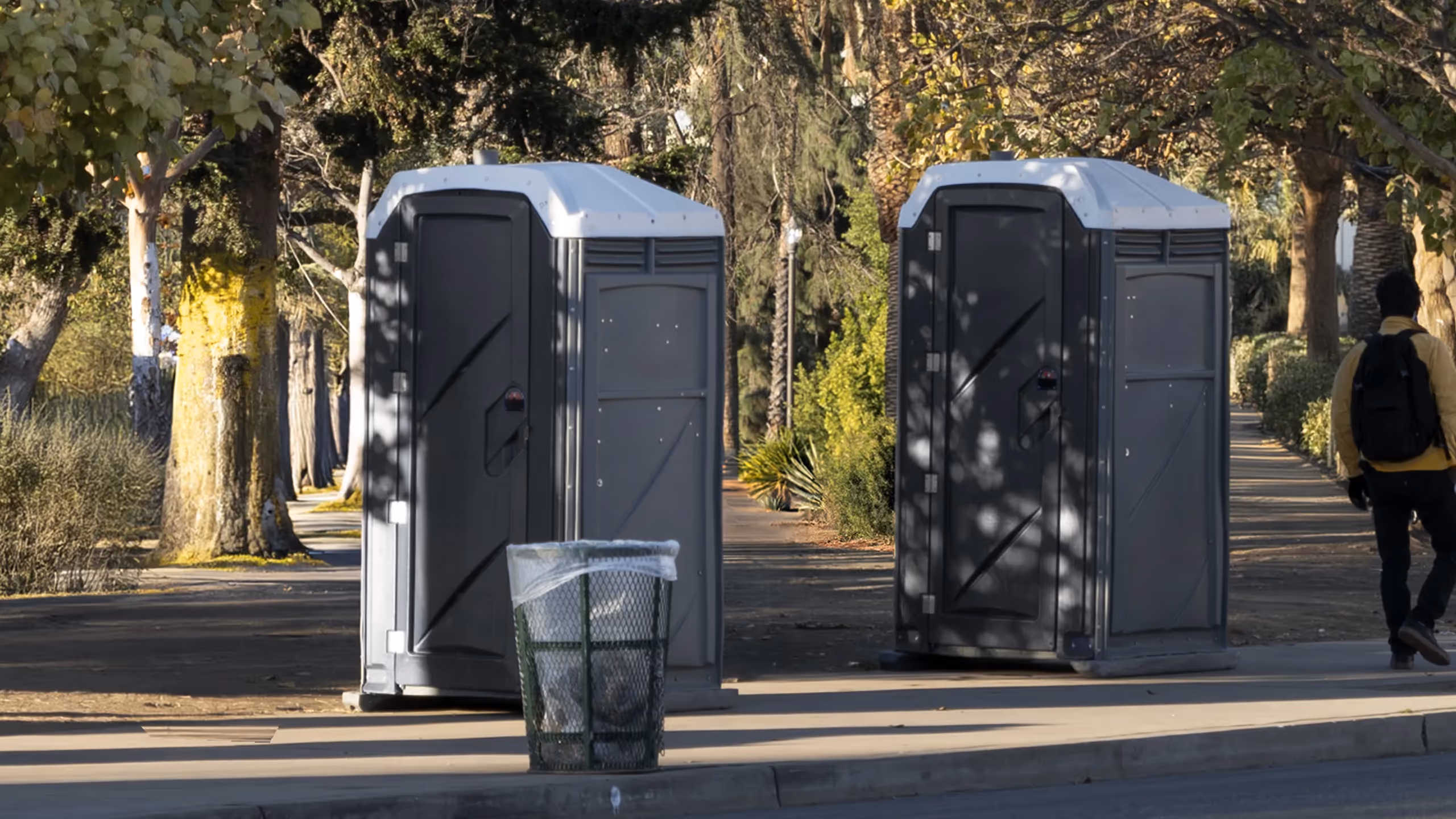 Two portable restrooms positioned along a sidewalk, with a trash bin in the foreground.