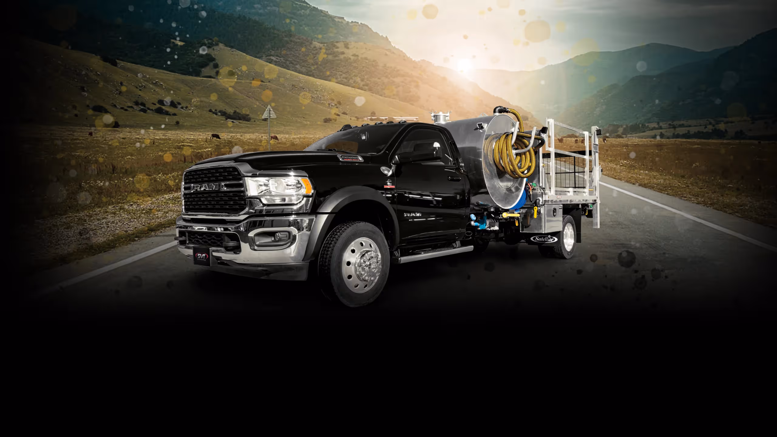 Black Ram truck with industrial tank and yellow hose on highway with mountainous landscape.