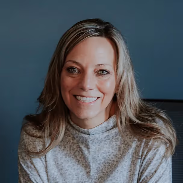 Smiling woman with long blonde hair in a light gray patterned sweater against a blue background.
