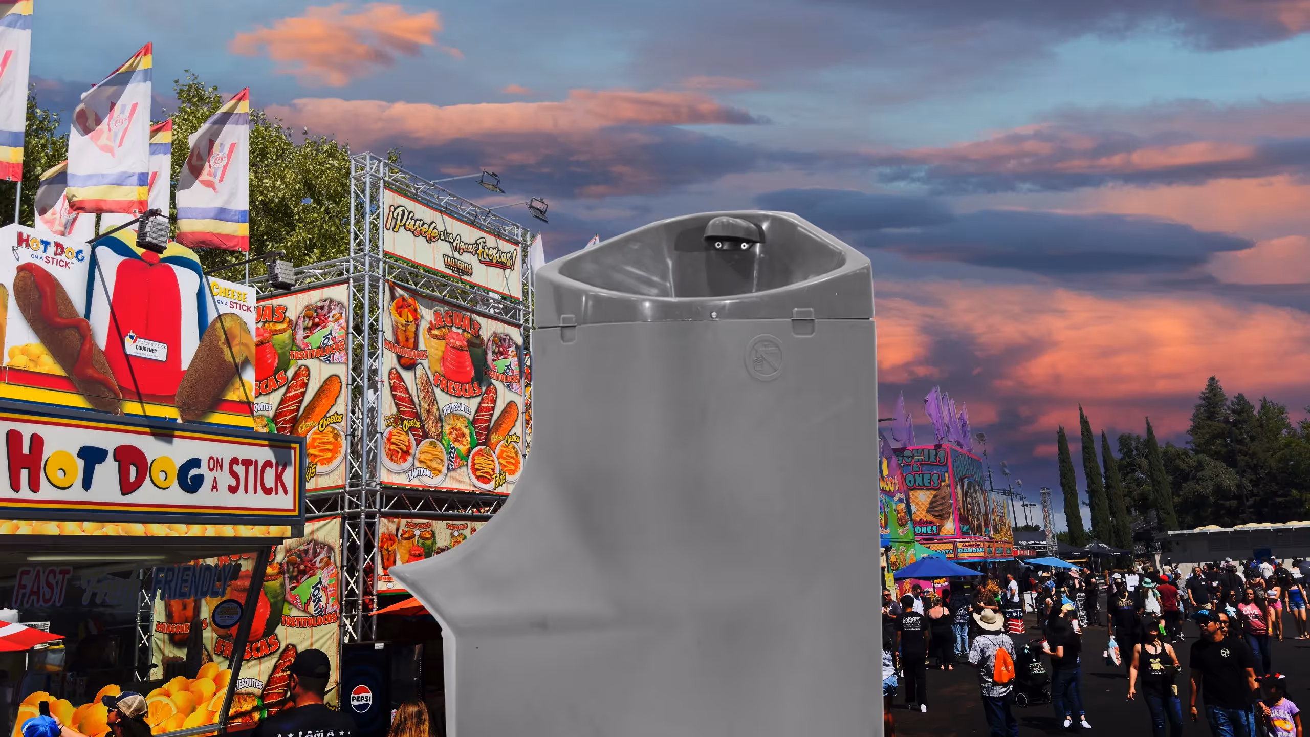 Handwash station in foreground at a crowded outdoor fair with colorful food stalls and sunset sky.