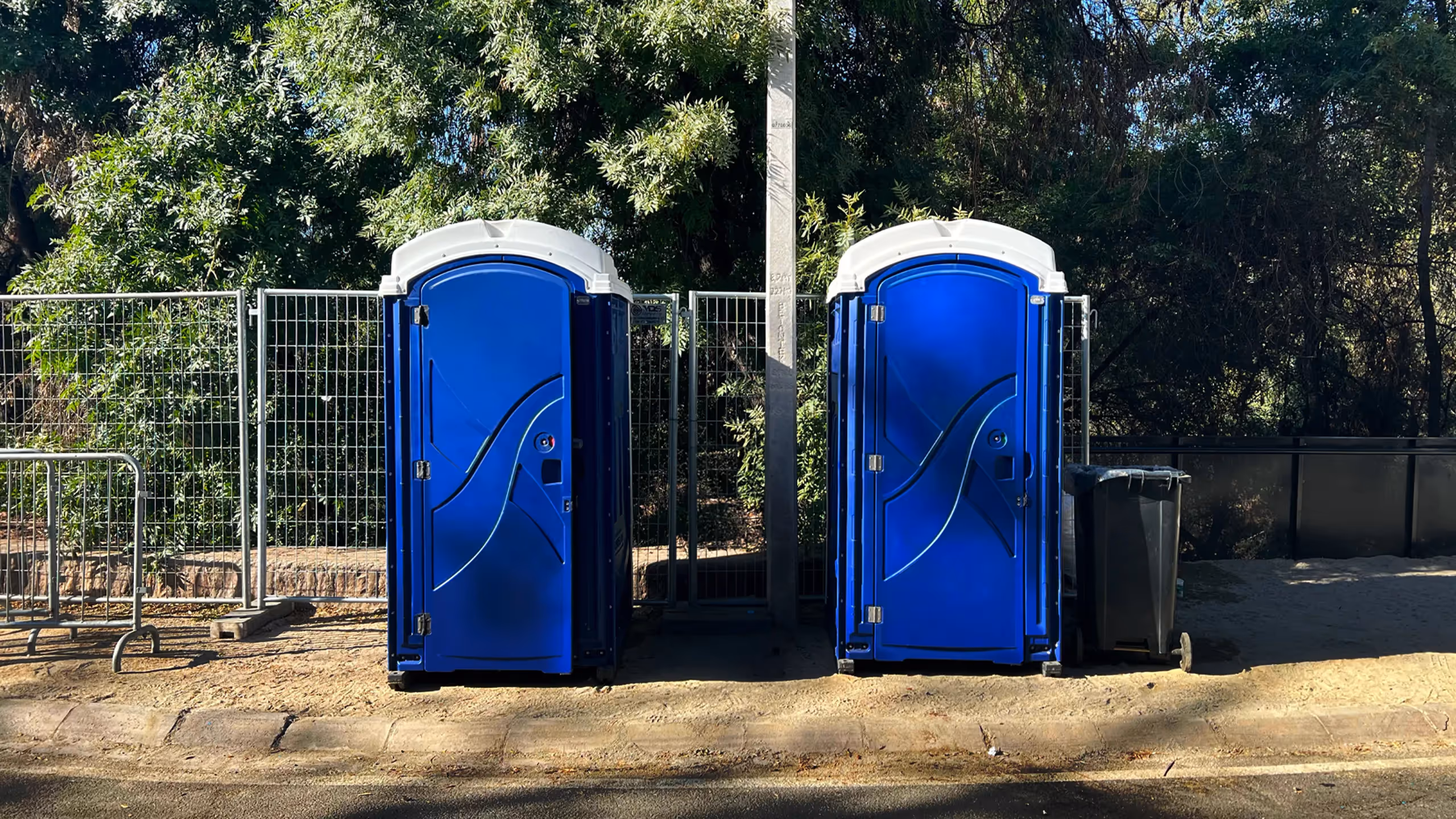 Two blue portable restrooms placed next to a fenced area with a trash bin nearby.