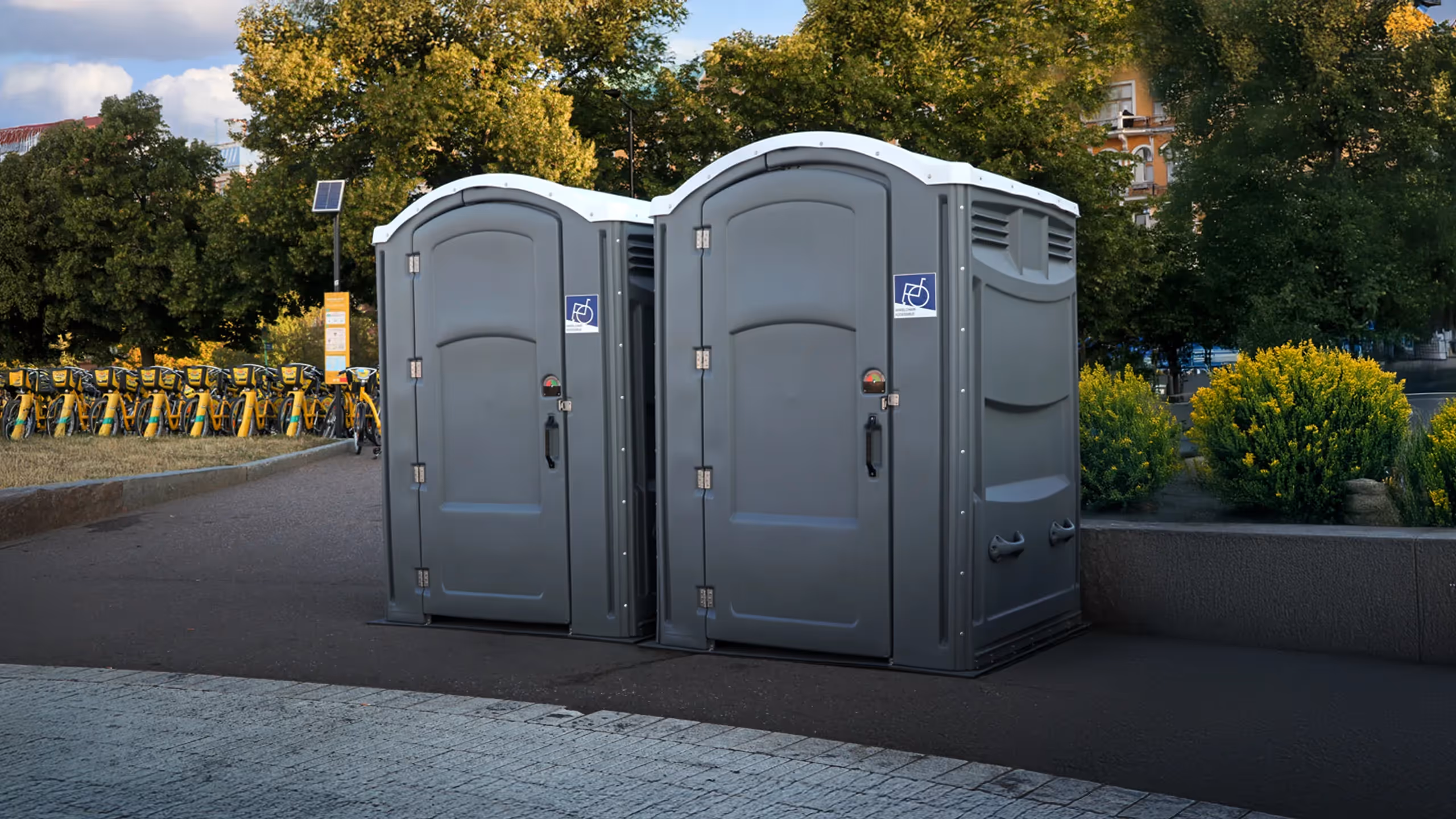 Two portable toilets placed next to a pathway, surrounded by greenery and bike racks in the