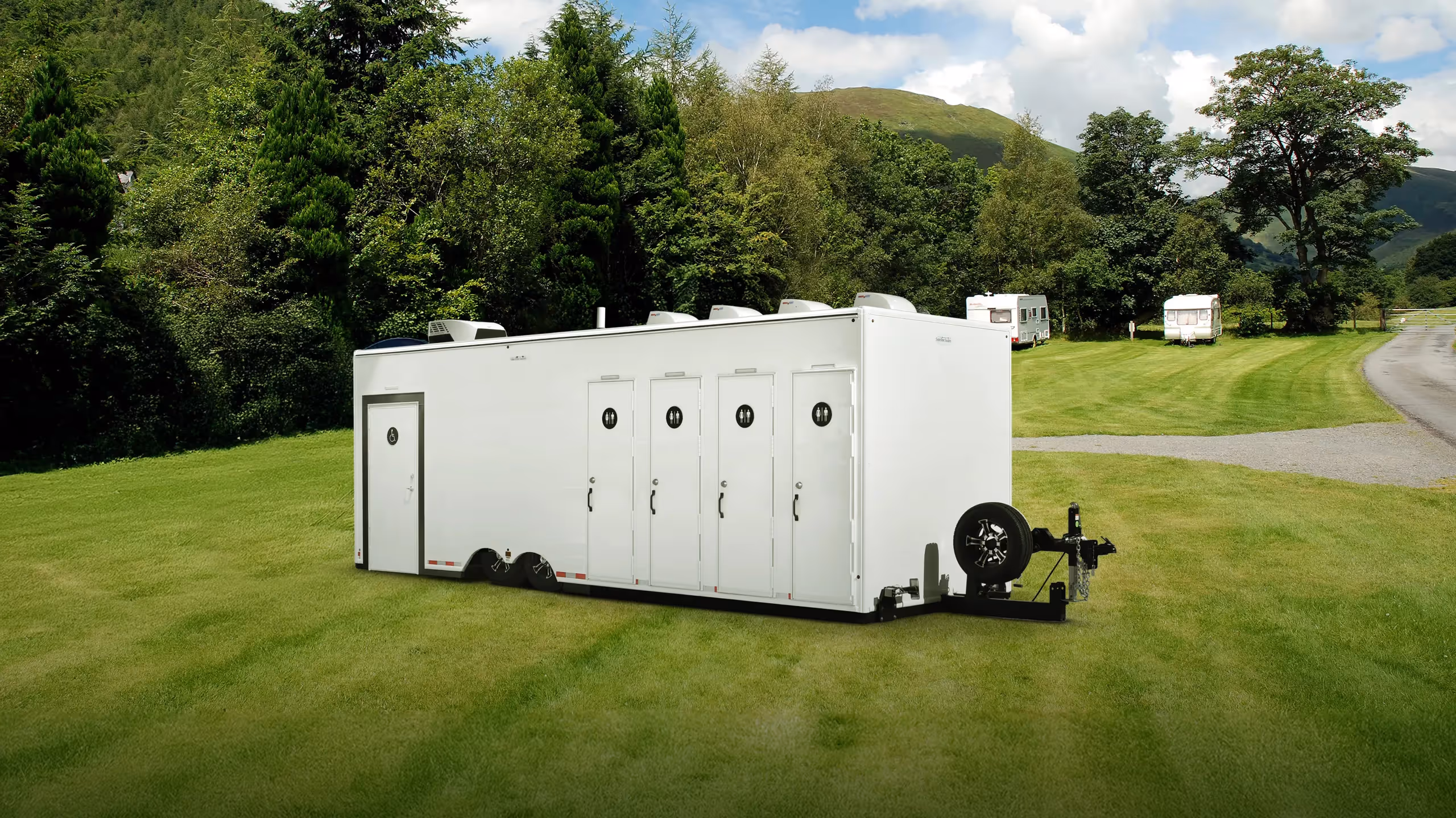 A large white trailer with four restroom doors, set on a grassy area surrounded by trees.