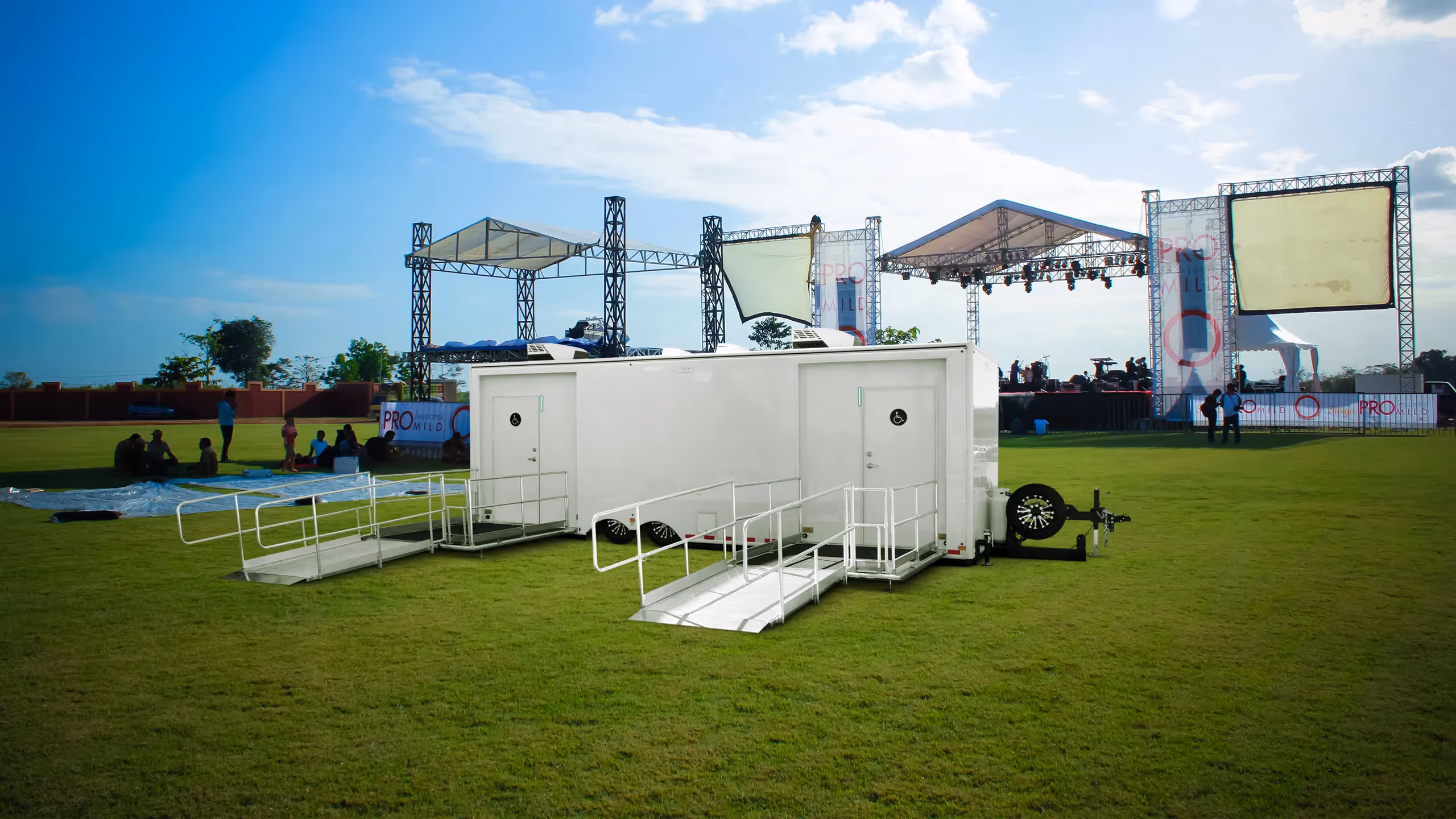 Two mobile restroom trailers sit on green grass, with a stage in the background and blue sky above.