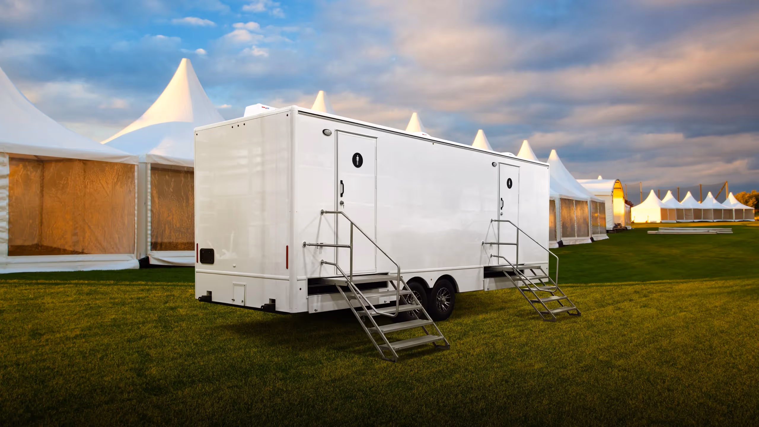 White 10-station restroom trailer parked on grass, with large tents in the background under a cloudy