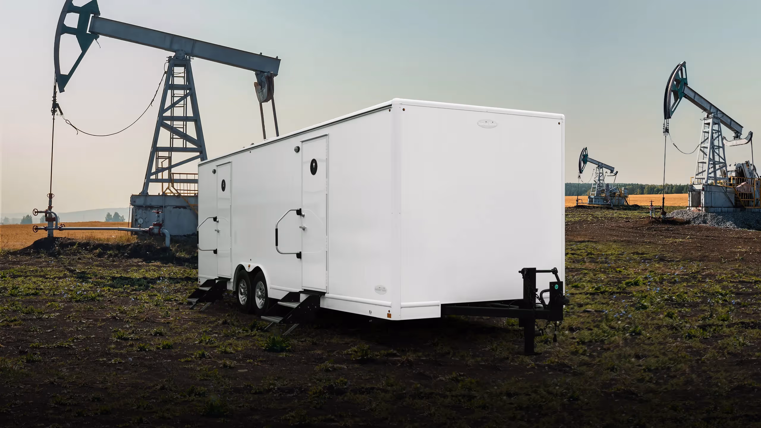 A white 8x26' trailer in an oil field with drilling rigs in the background.