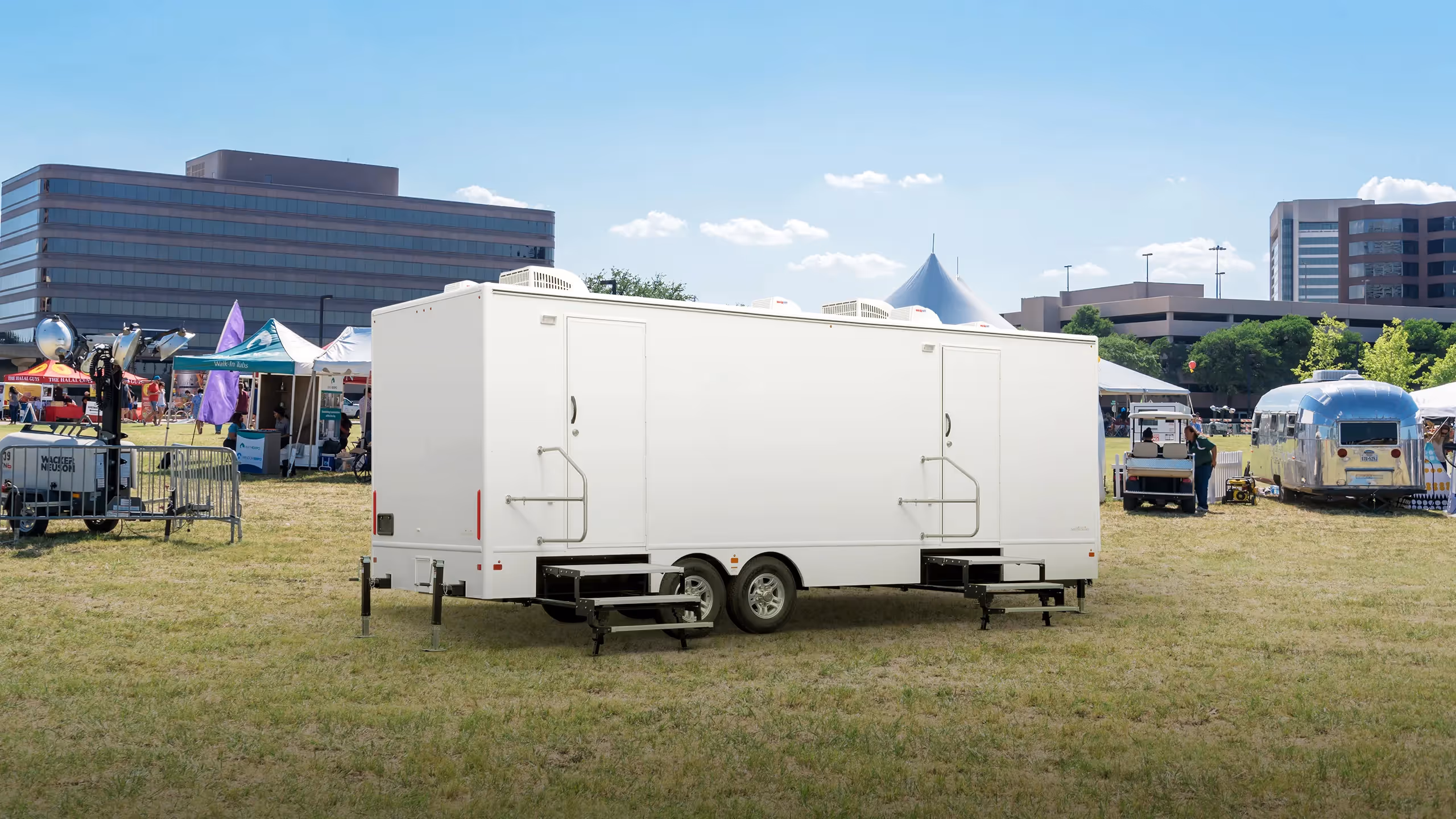 White 8-station restroom trailer on grassy field with buildings and tents in the background.