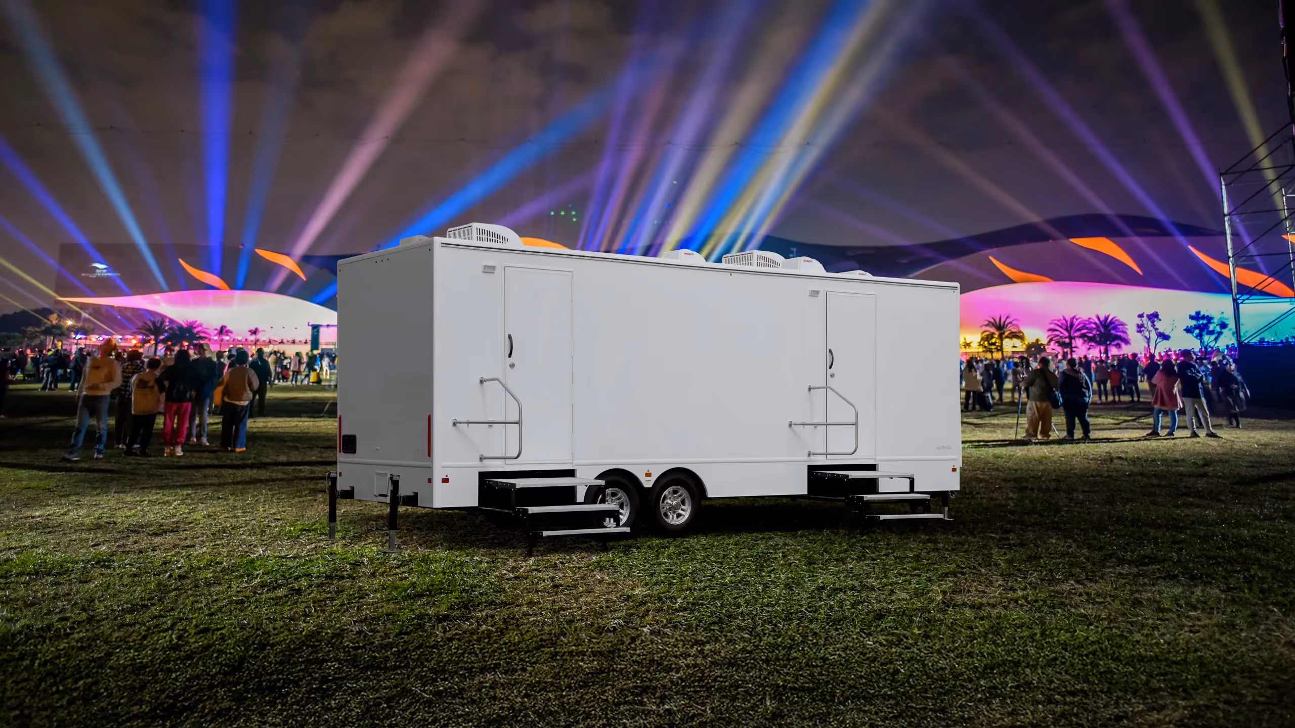 A white 10-station restroom trailer positioned on grass at a nighttime event with colorful lights.
