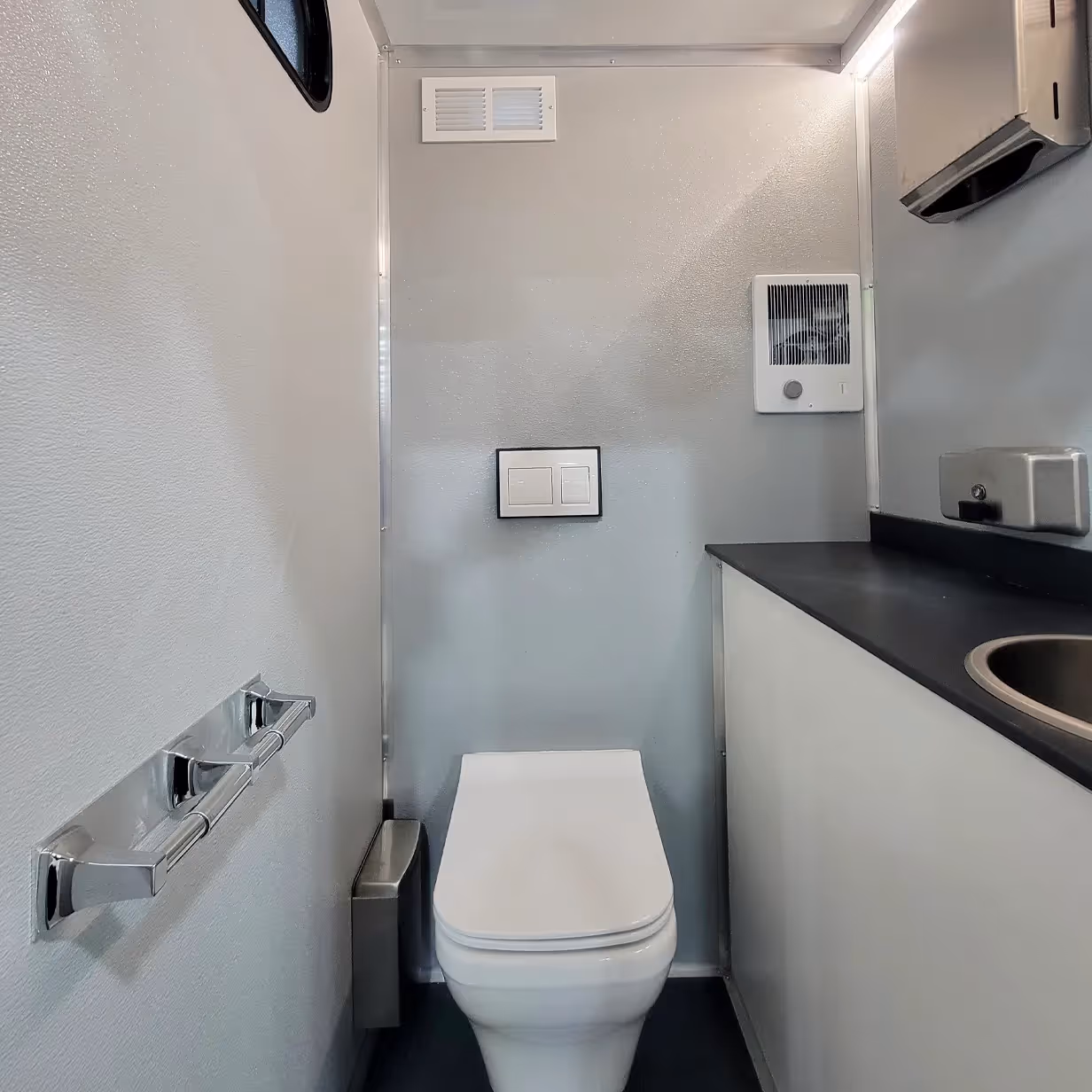 Compact restroom with a wall-mounted toilet, ventilation, and a countertop with a sink.