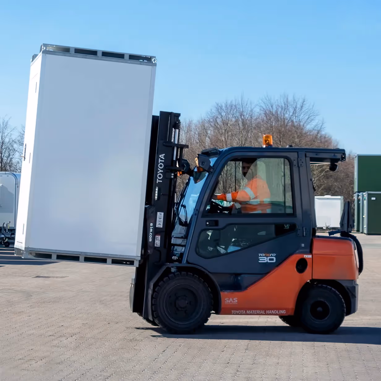 Forklift lifting a large white container outdoors with driver in high-visibility jacket.