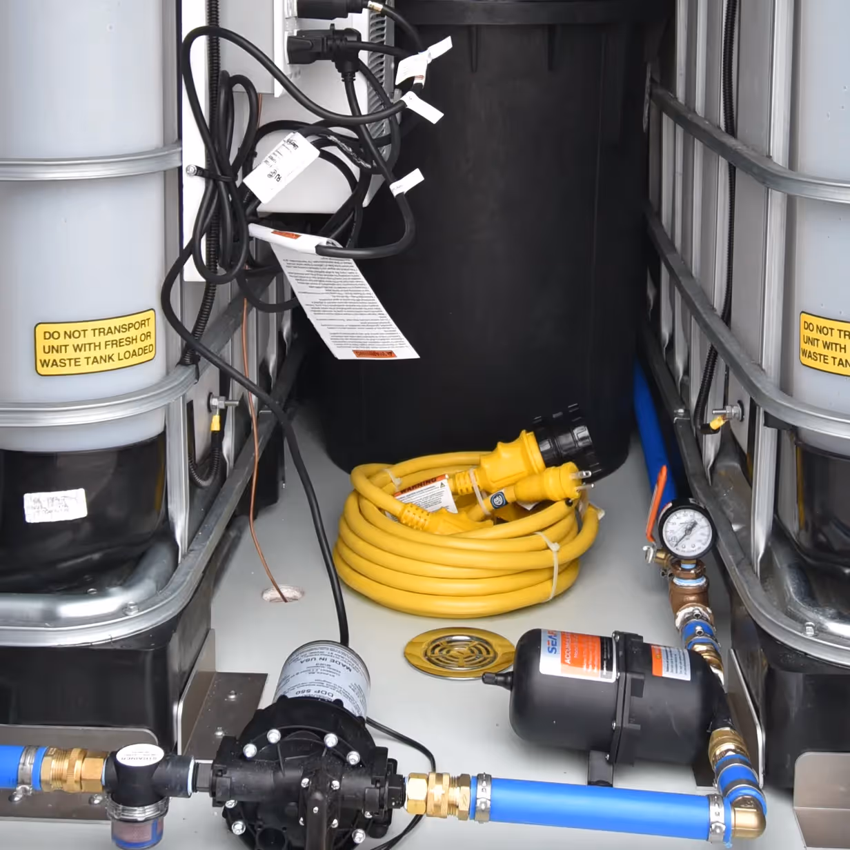 Control compartment with coiled yellow cable, pressure gauge, black pump, and two large white tanks with warning labels.