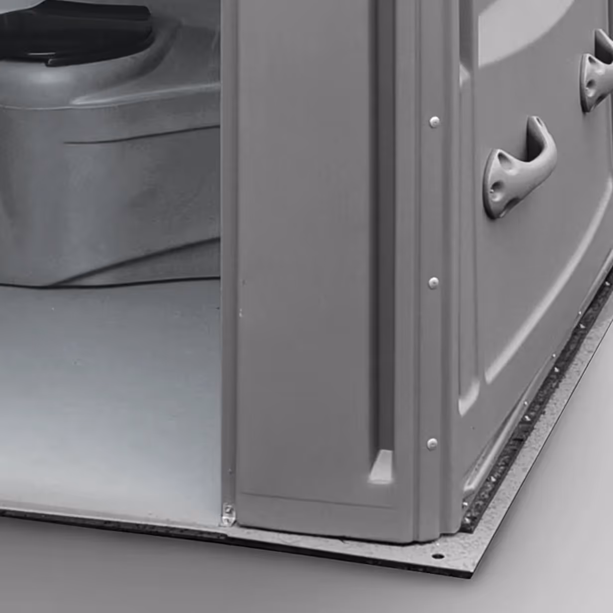 Close-up of a gray portable toilet corner showing the base plate and part of the door and wall.