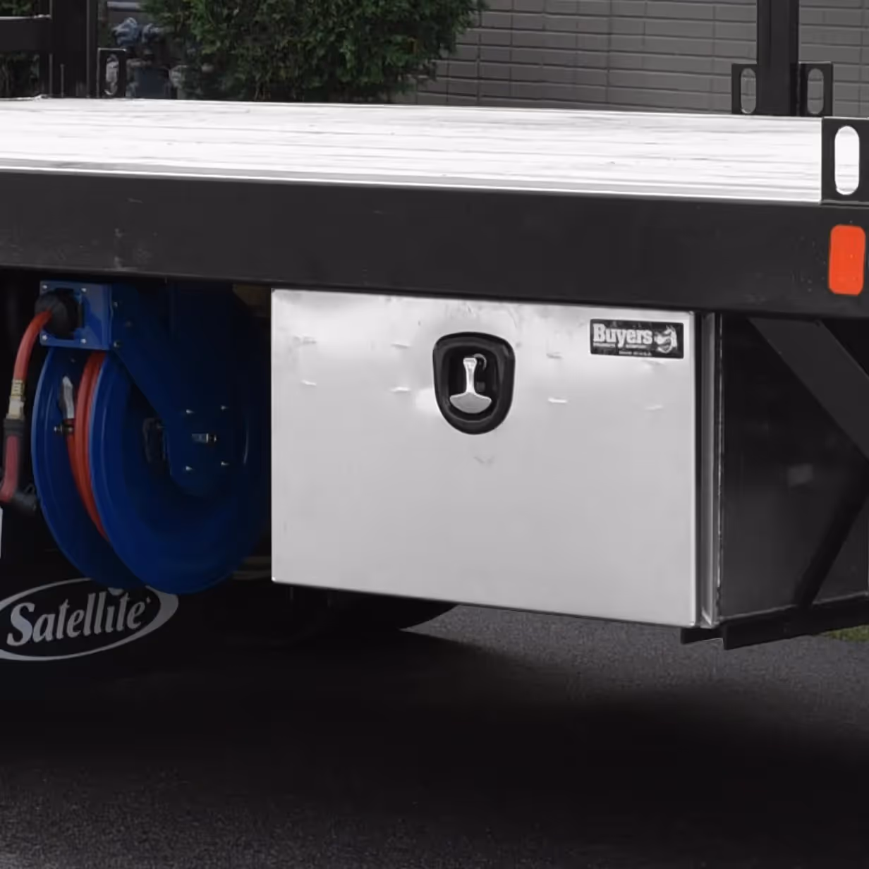 Stainless steel equipment storage box mounted under flatbed truck with blue hose reel beside it.