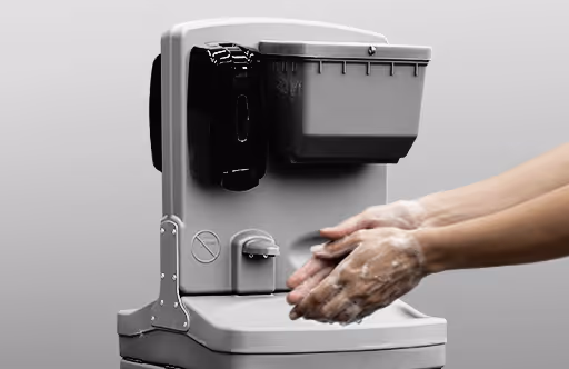 Hands are being washed under a soap dispenser with a gray container in the background.