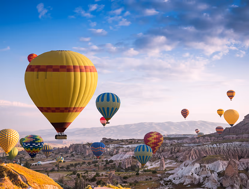 Colorful hot air balloons float over rocky landscapes under a blue sky with scattered clouds.