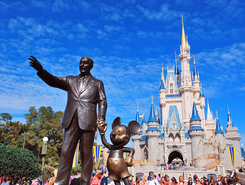 Bronze statue of a man and Mickey Mouse in front of a fairytale castle and blue sky.
