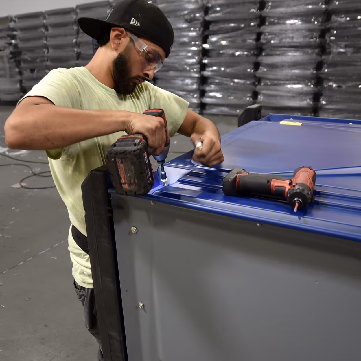 A man with glasses and a cap works on assembling a metal structure using tools in a warehouse.