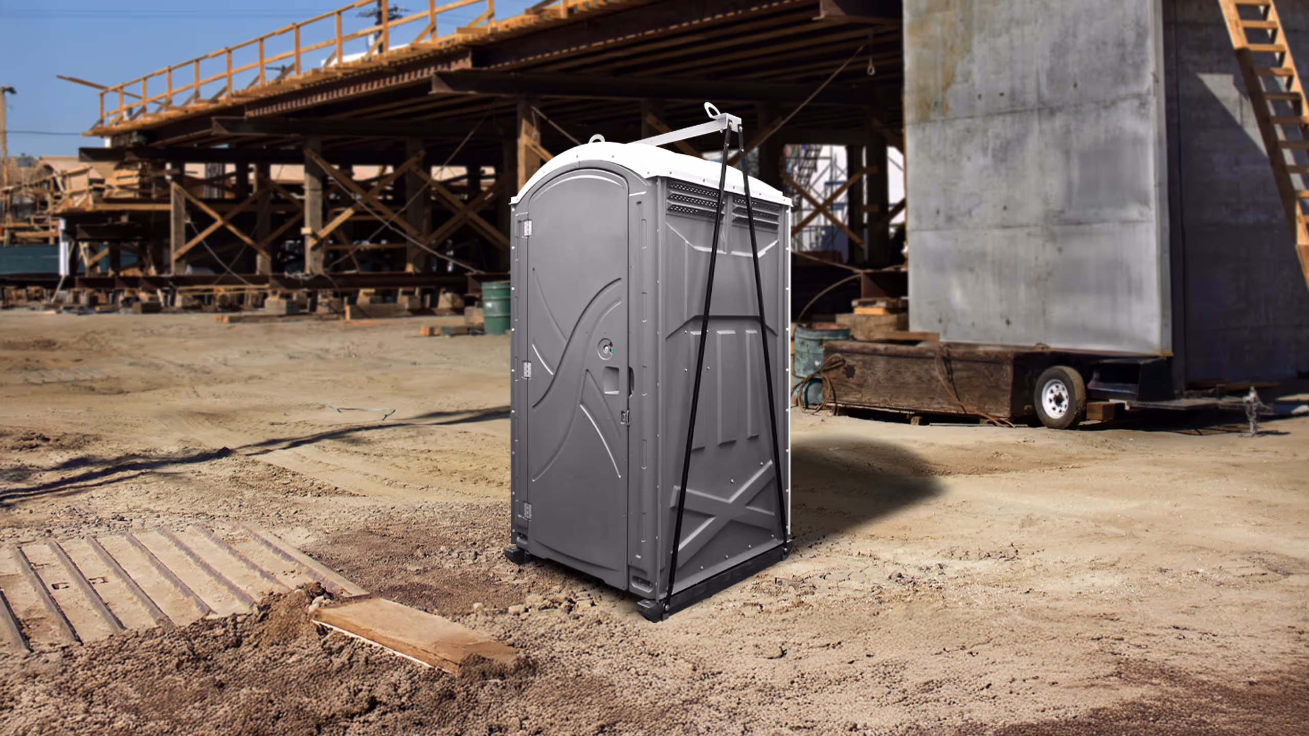 Gray portable toilet secured with black straps on a construction site with scaffolding and dirt ground.