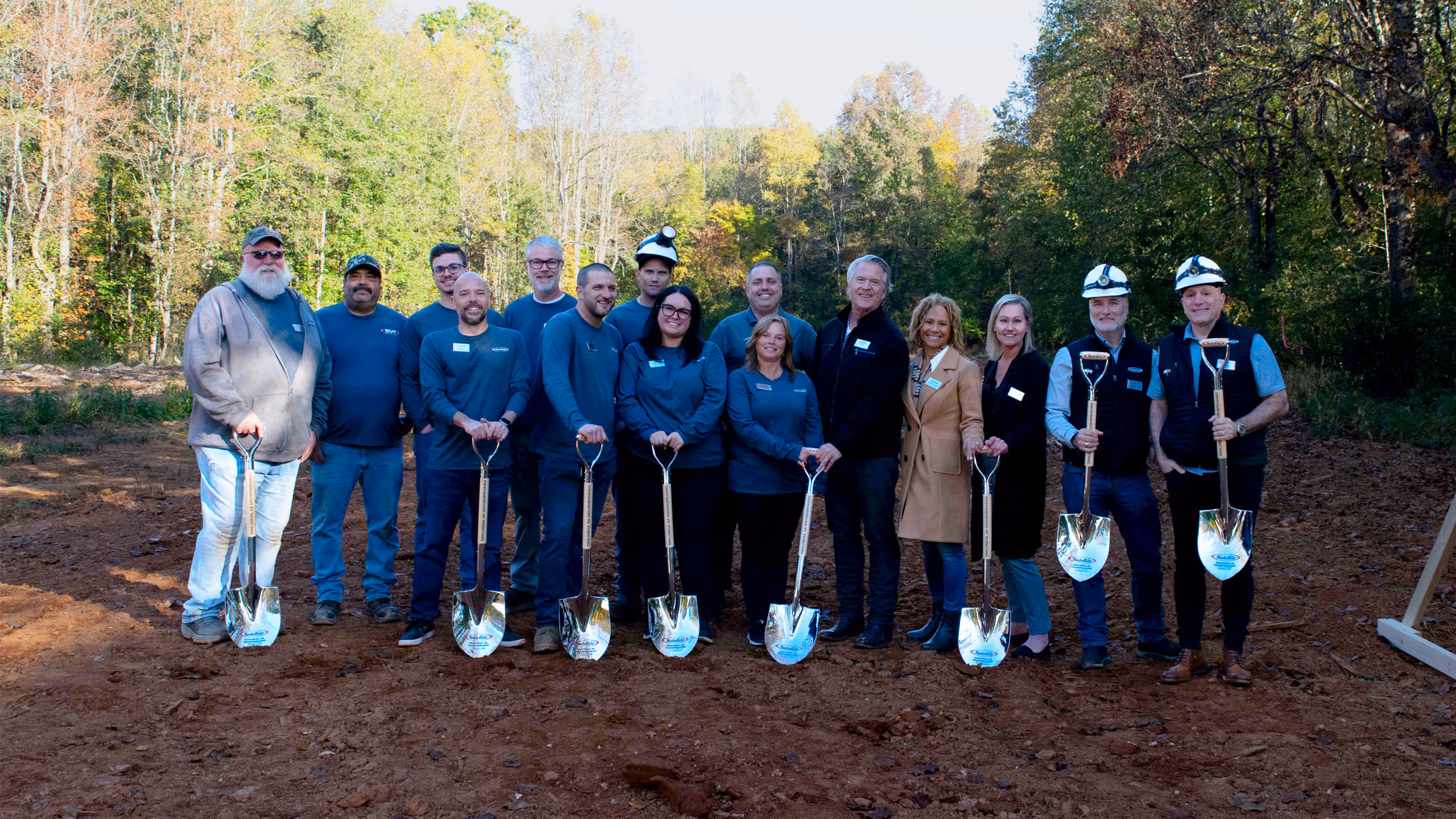 Group of people in work attire holding shovels at a groundbreaking event in a wooded area.