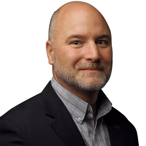 Bald man with grey beard wearing a checked shirt and dark blazer smiling against black background.