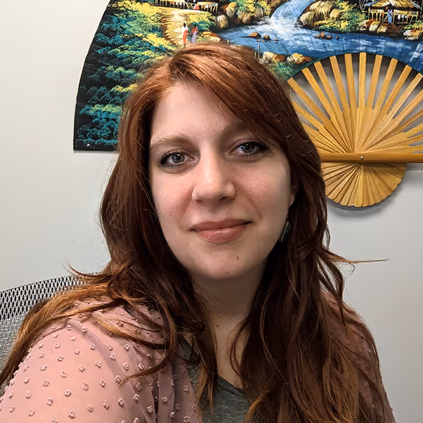Woman with long red hair and pink blouse sits in front of a decorative fan and painting on wall.