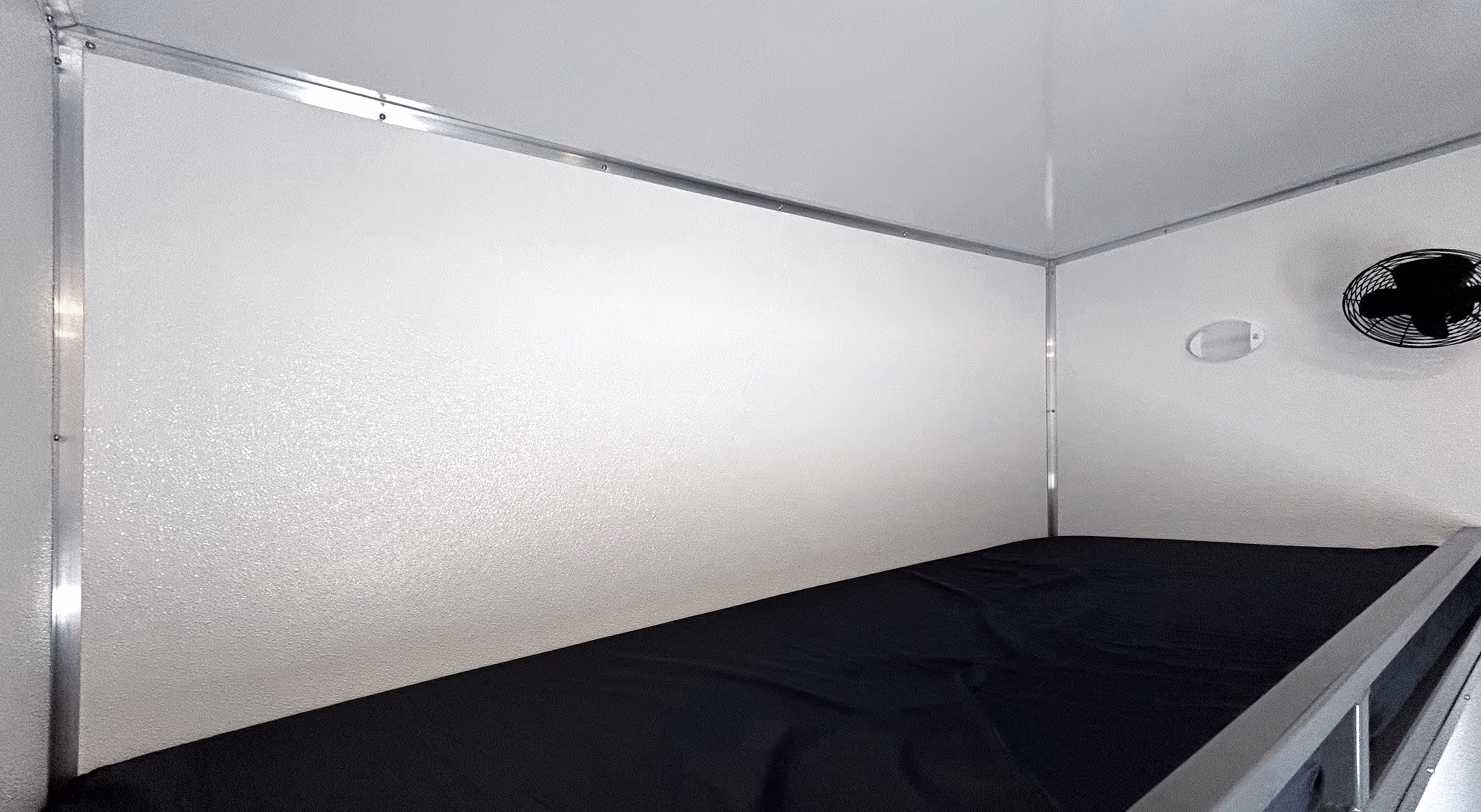 White-paneled interior with a black mattress on a bunk bed and a small fan mounted on the wall.