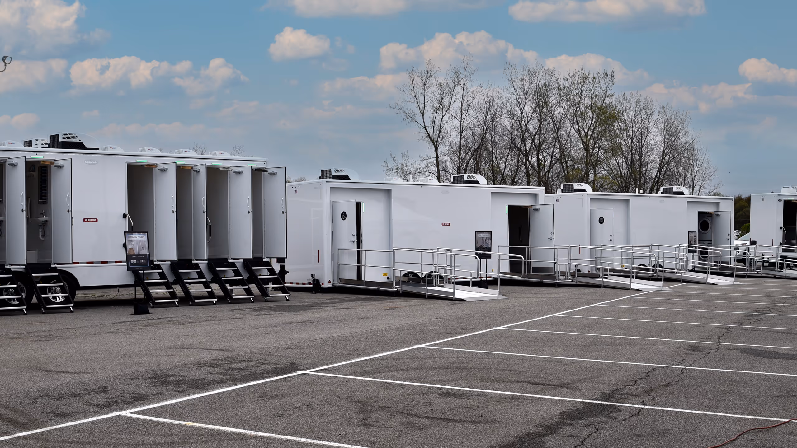 Row of portable restrooms and hygiene trailers in a parking lot under a cloudy sky.