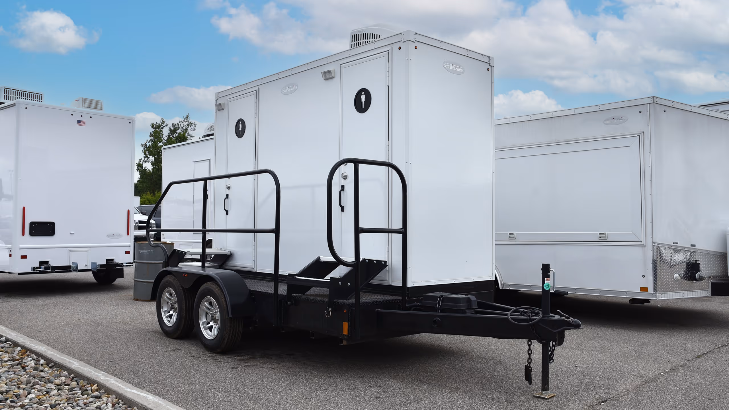 A portable restroom trailer with black wheels and a tow hitch, parked among white trailers.