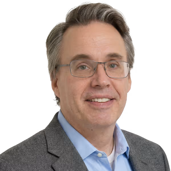 Middle-aged man with glasses smiling, wearing a gray blazer over a blue collared shirt.