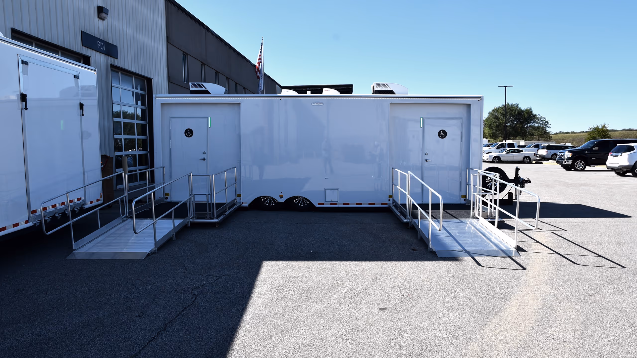 Two white trailers with accessible ramps are parked beside a building on a sunny day.