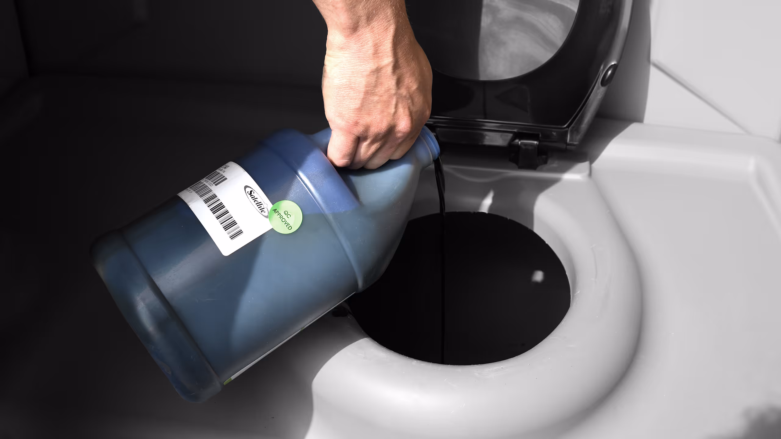 A hand pours a blue liquid from a container into a black opening, possibly a waste tank.
