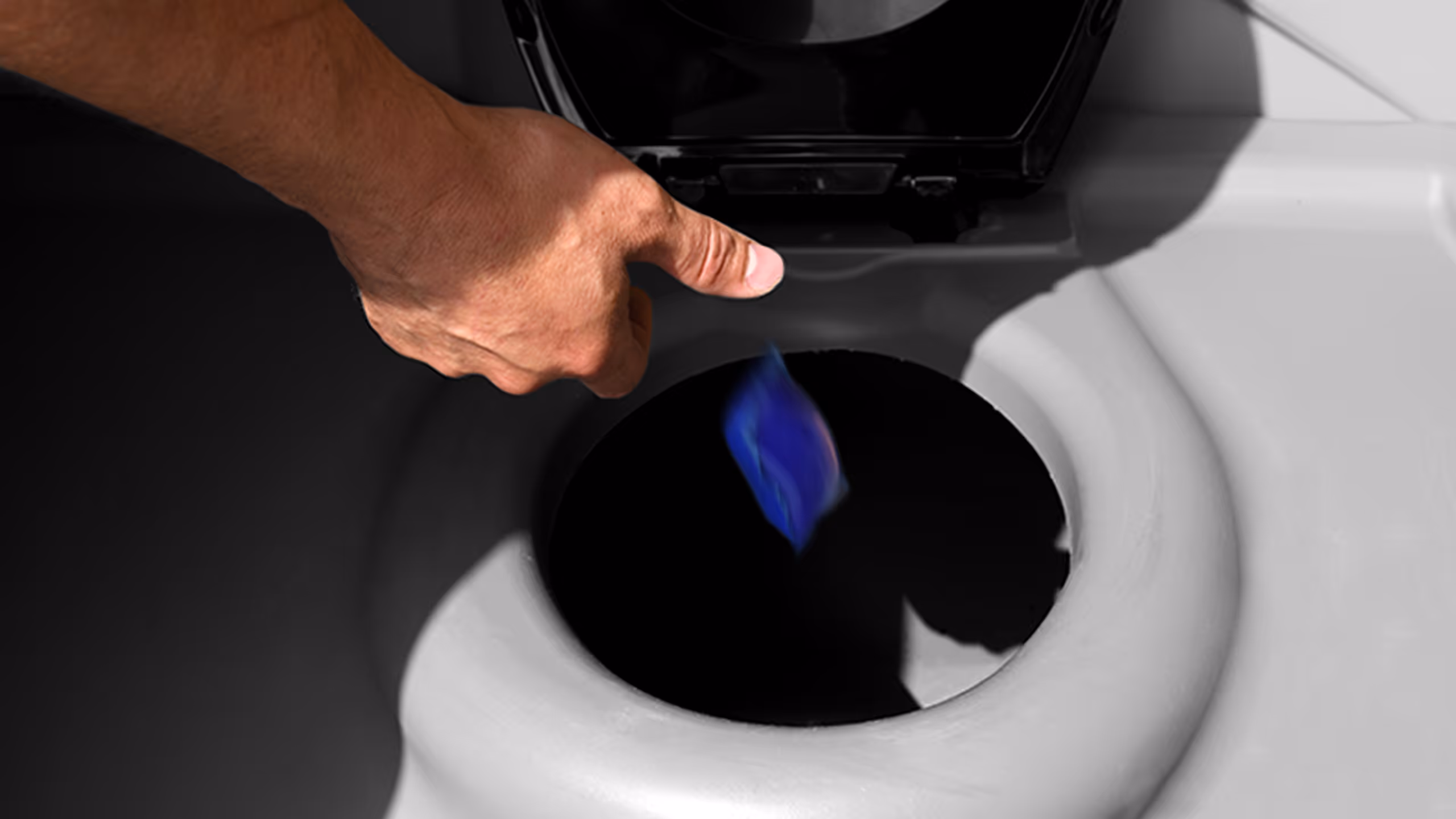A hand is dropping a blue object into a toilet bowl from above, with shadows in the background.
