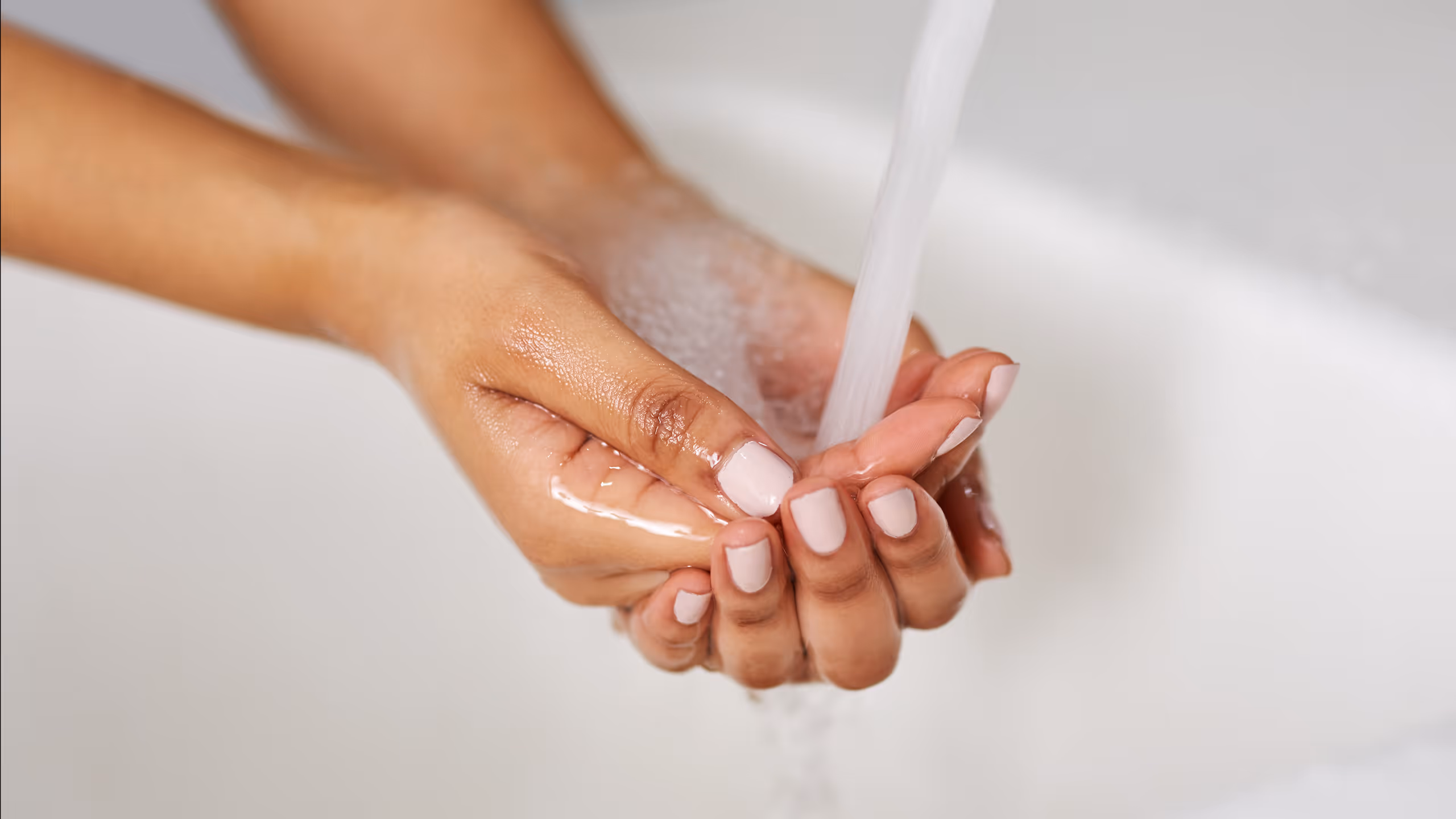 Hands with light pink nail polish are washing under running water in a sink.