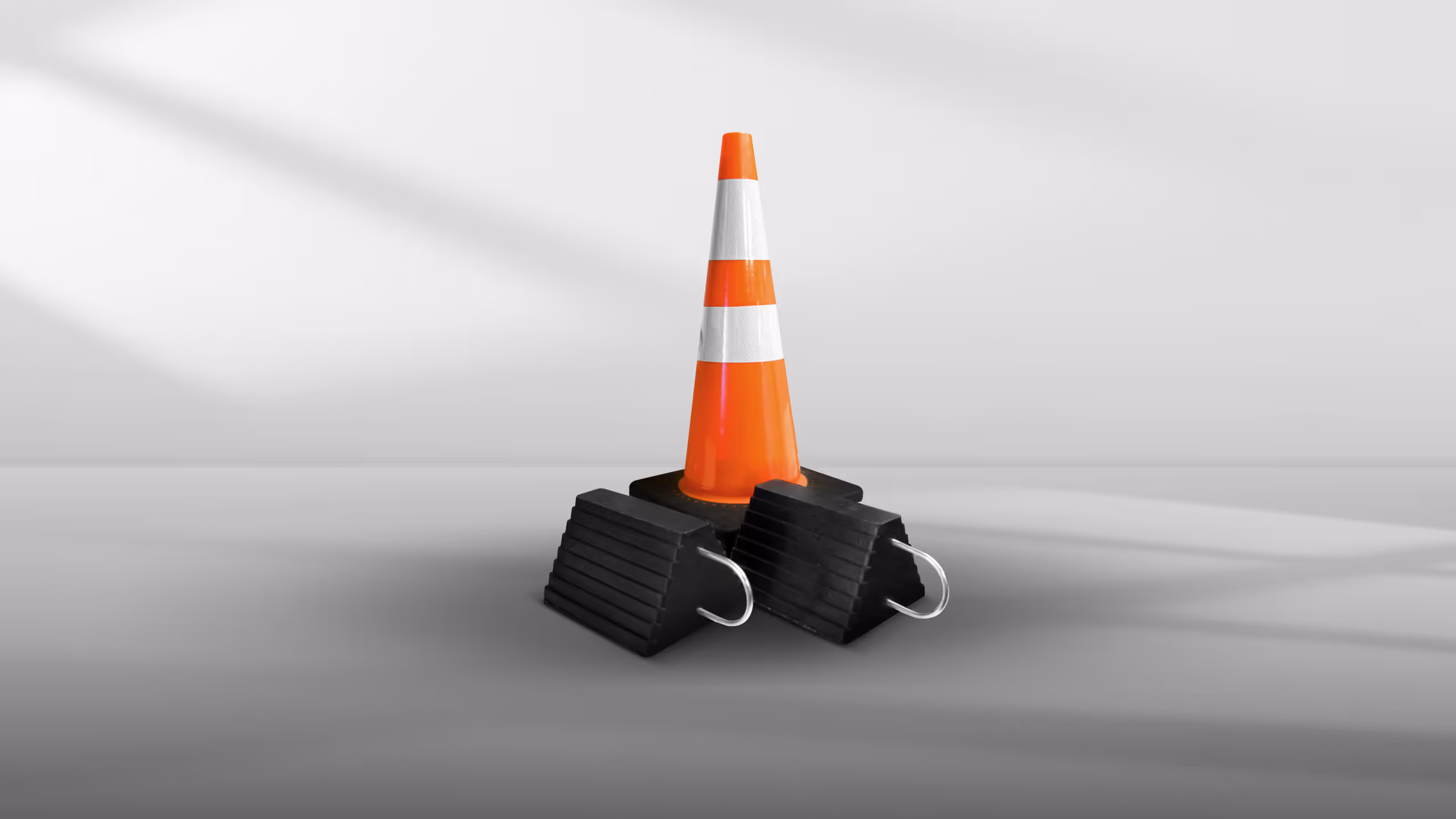 A traffic cone with white reflective stripes, positioned on two black rubber wheel chocks.
