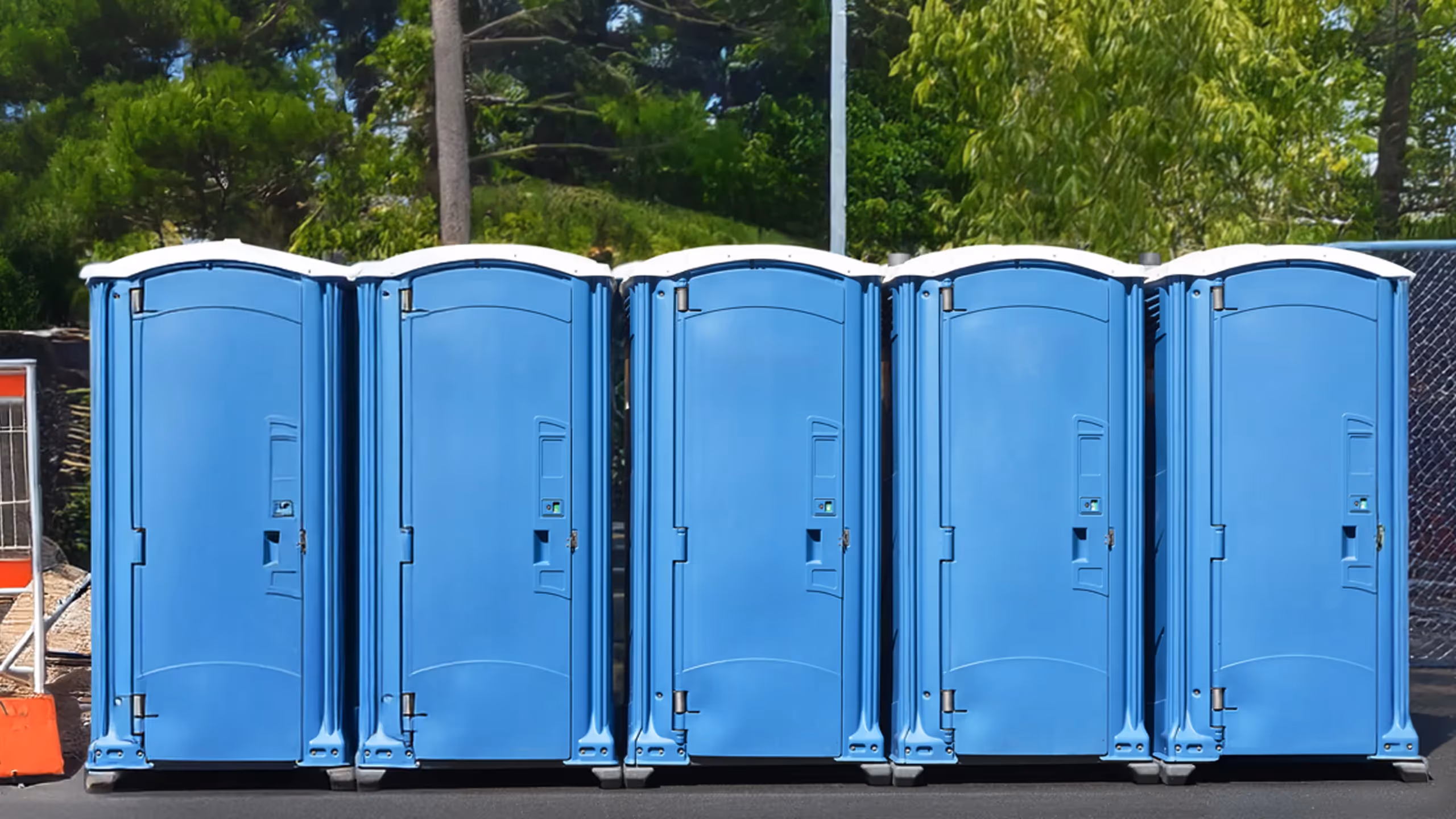 Five blue portable toilets lined up outdoors on a sunny day with trees in the background.