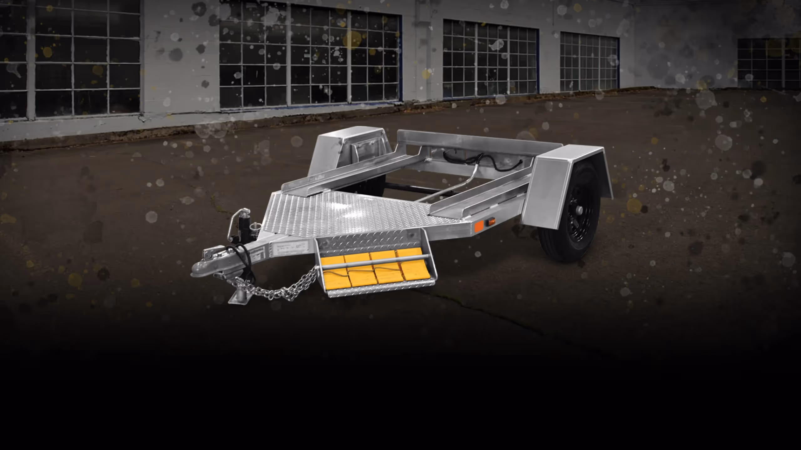 Metal trailer frame with one wheel and yellow blocks in front, set on dark pavement.