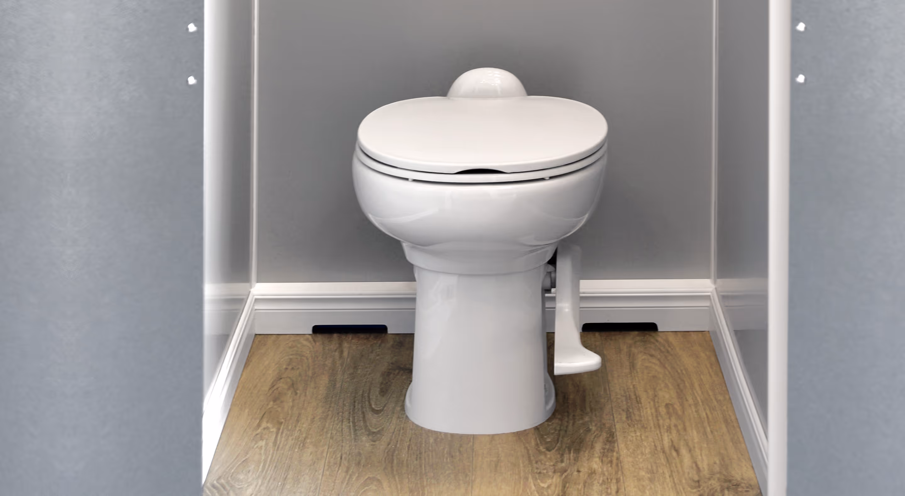 White toilet with closed lid and pedal flush on wood floor between gray walls.