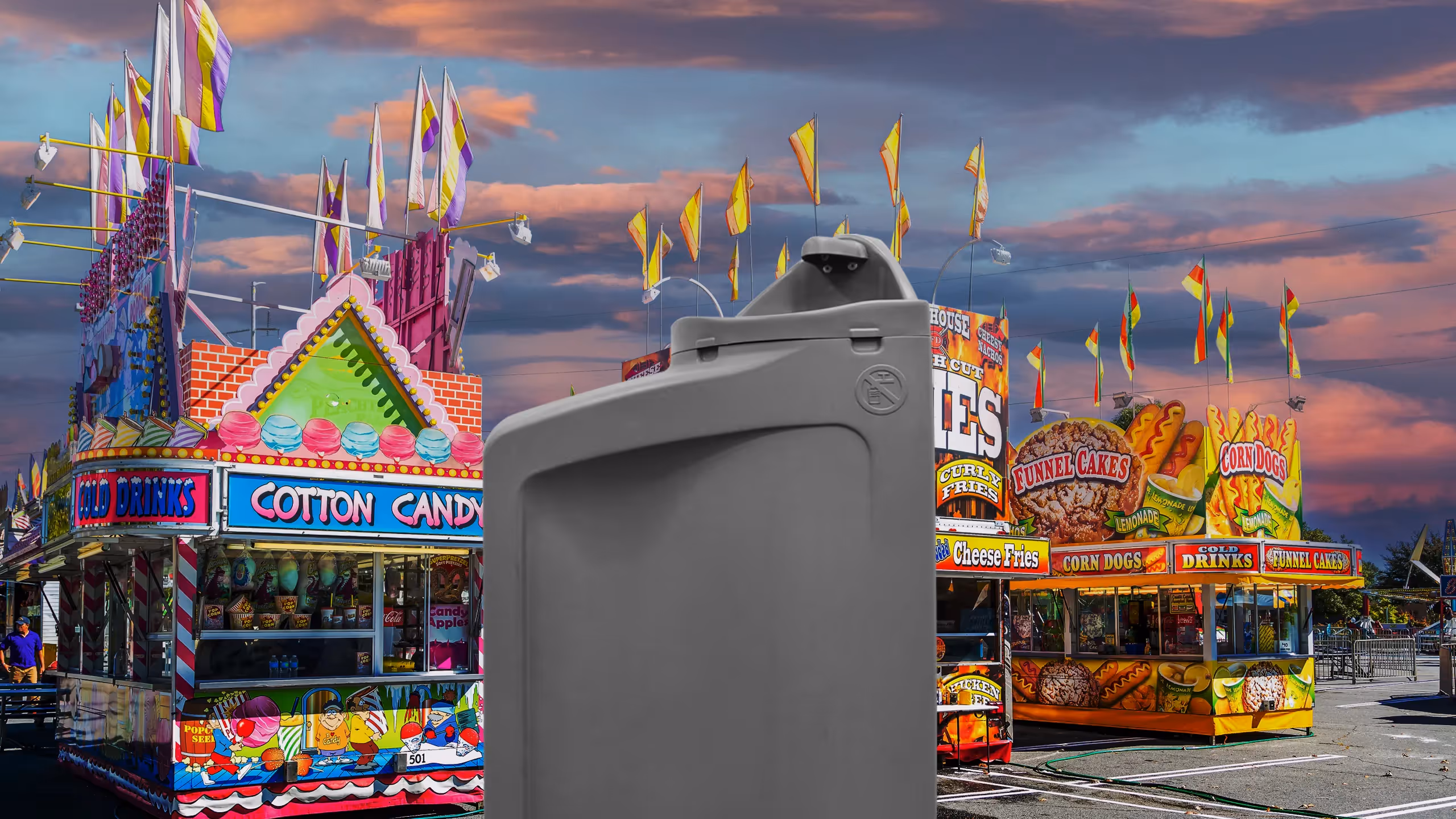 A gray container stands in front of brightly colored carnival food stalls against a sunset sky.