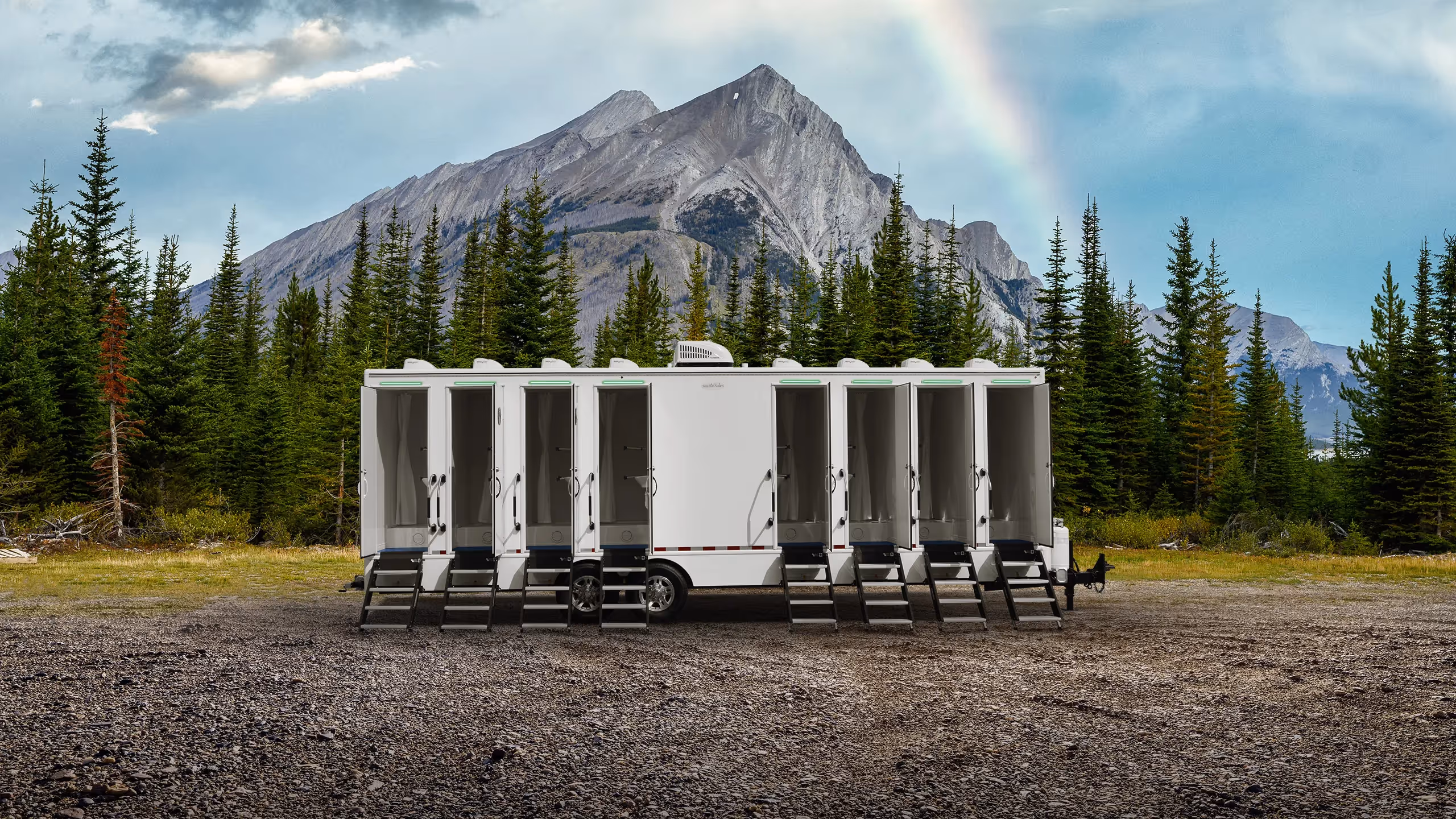 White mobile shower trailer with multiple open stalls in forest clearing, mountains, and rainbow behind.