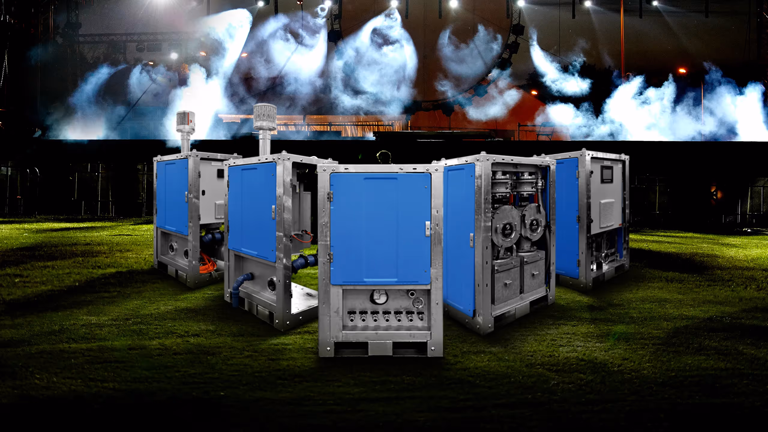 Five industrial machines with blue panels on grass with smoke and stage lights in the background.